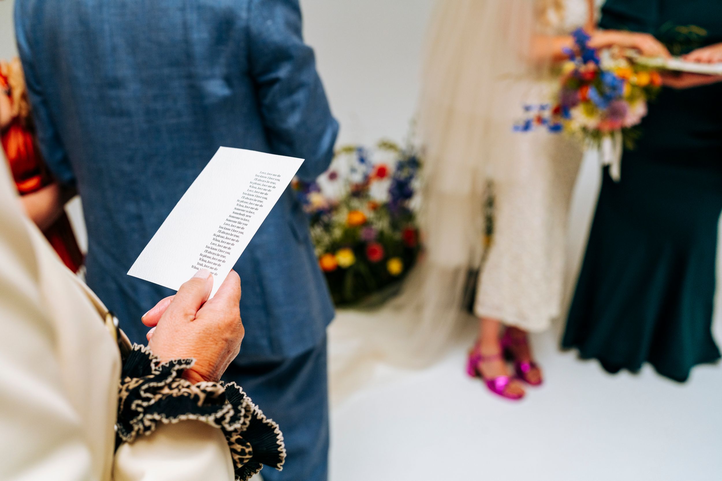 Wedding guest holds ceremony program while couple exchanges vows with colorful bouquet.