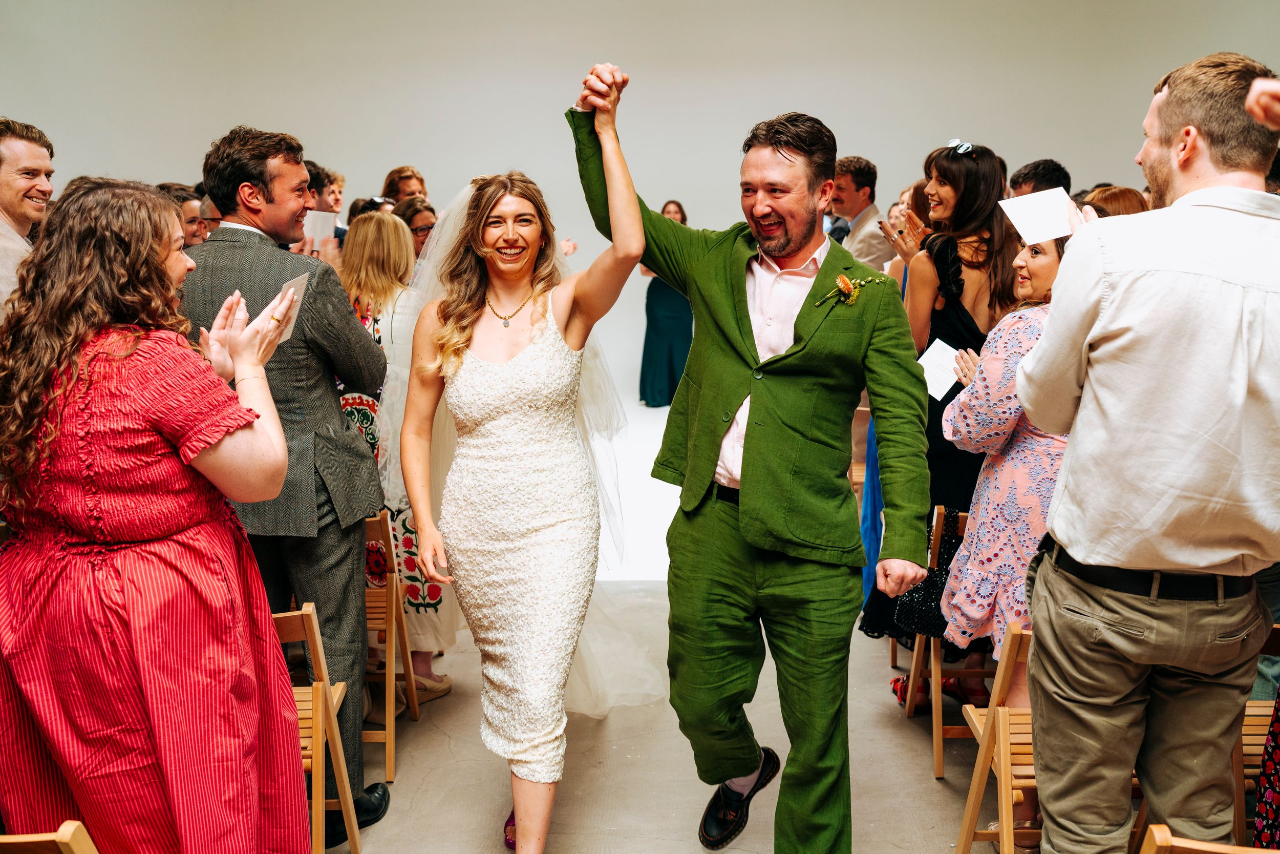 Bride in white dress and groom in green suit celebrate walking down the aisle after ceremony.
