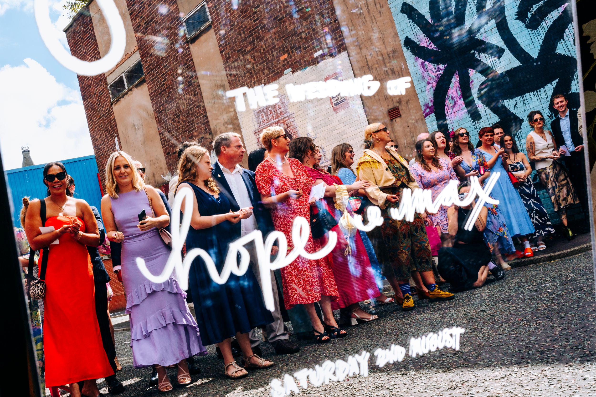 Wedding guests lined up outside a venue in front of a colorful hope mural on a sunny summer day.