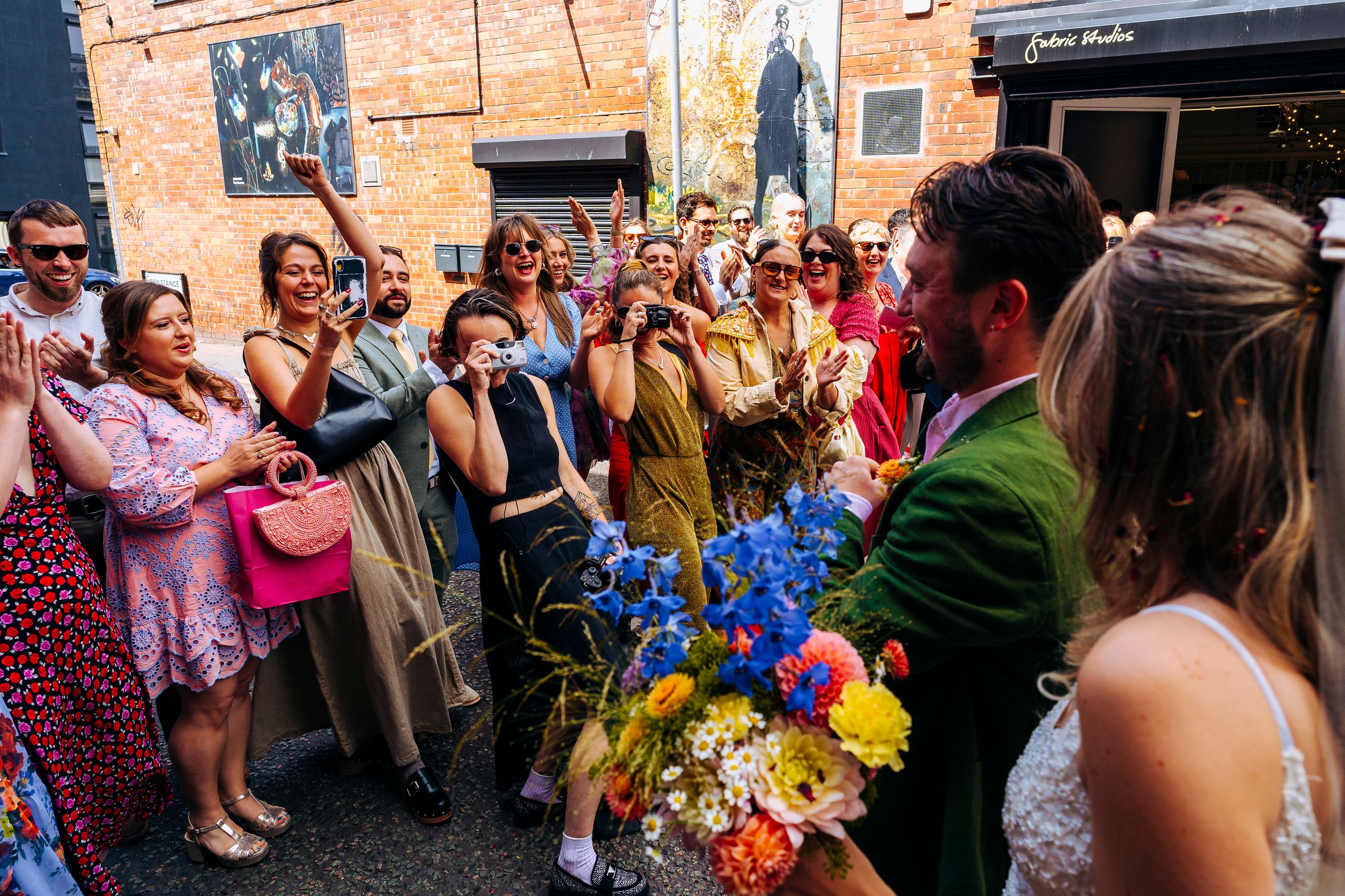 Wedding guests cheer as bride holds vibrant bouquet of blue and yellow flowers outside venue.