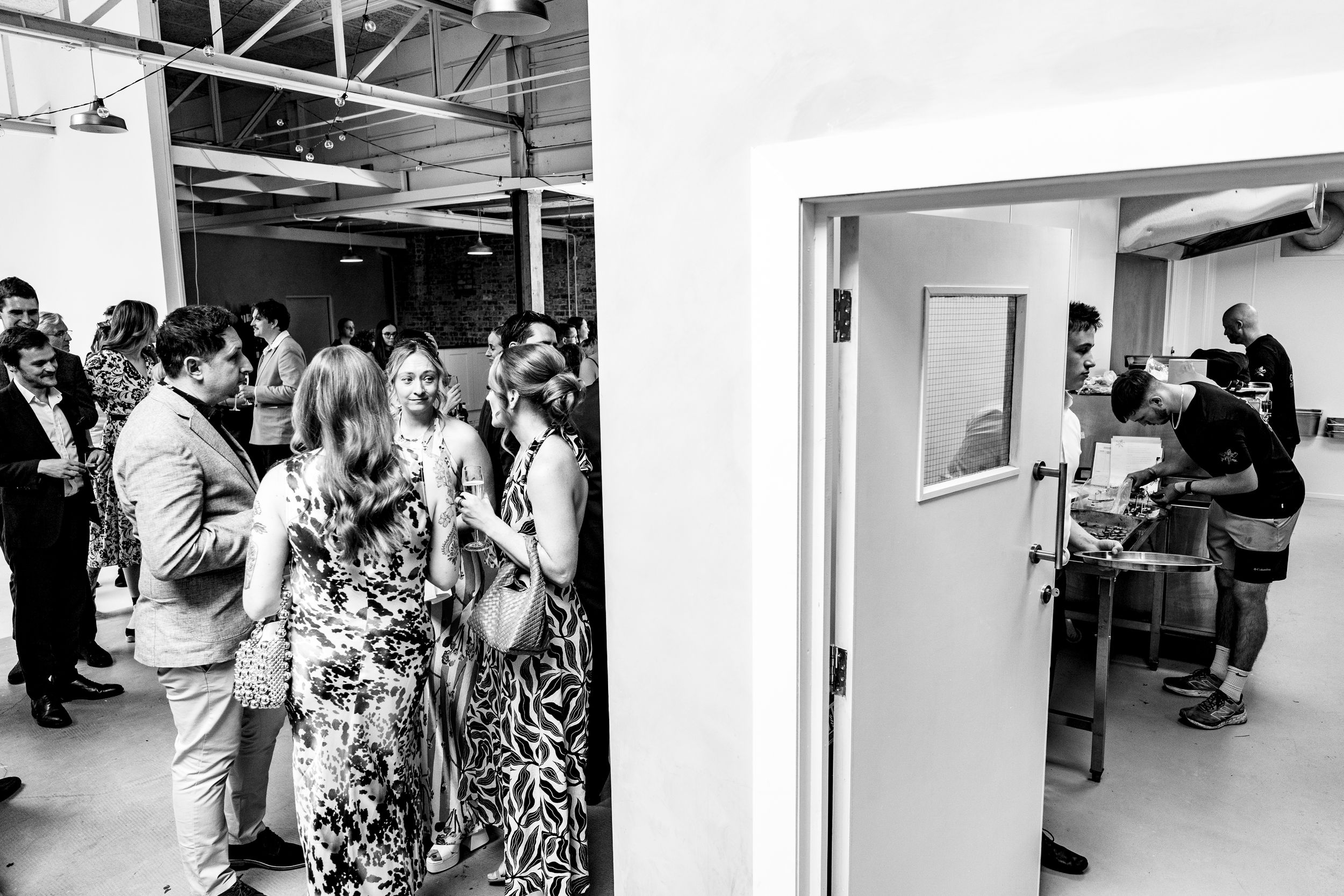 Black and white photo of wedding guests mingling in a modern industrial venue space.