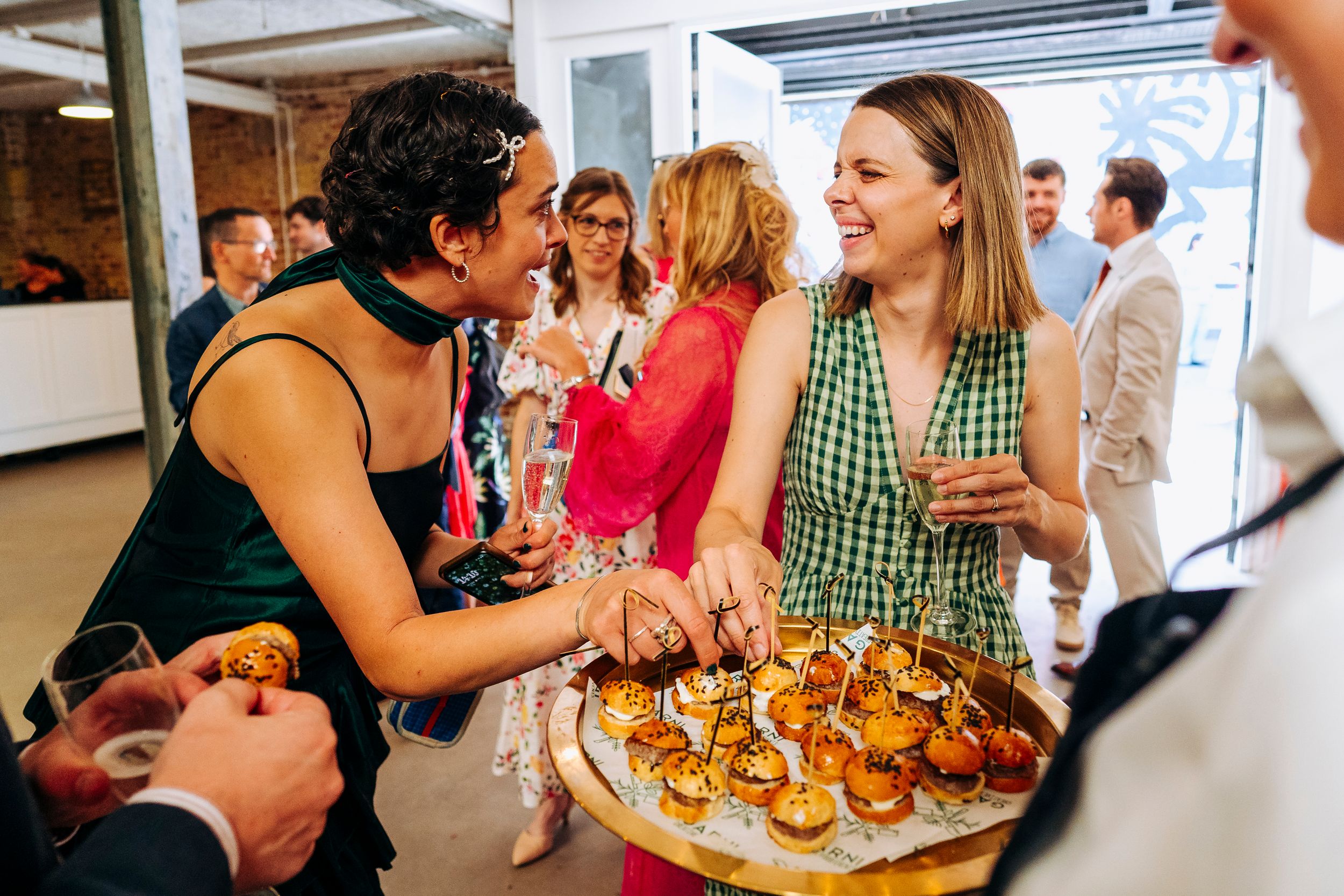Wedding guests smile and share canapes at a sunny indoor wedding reception celebration.