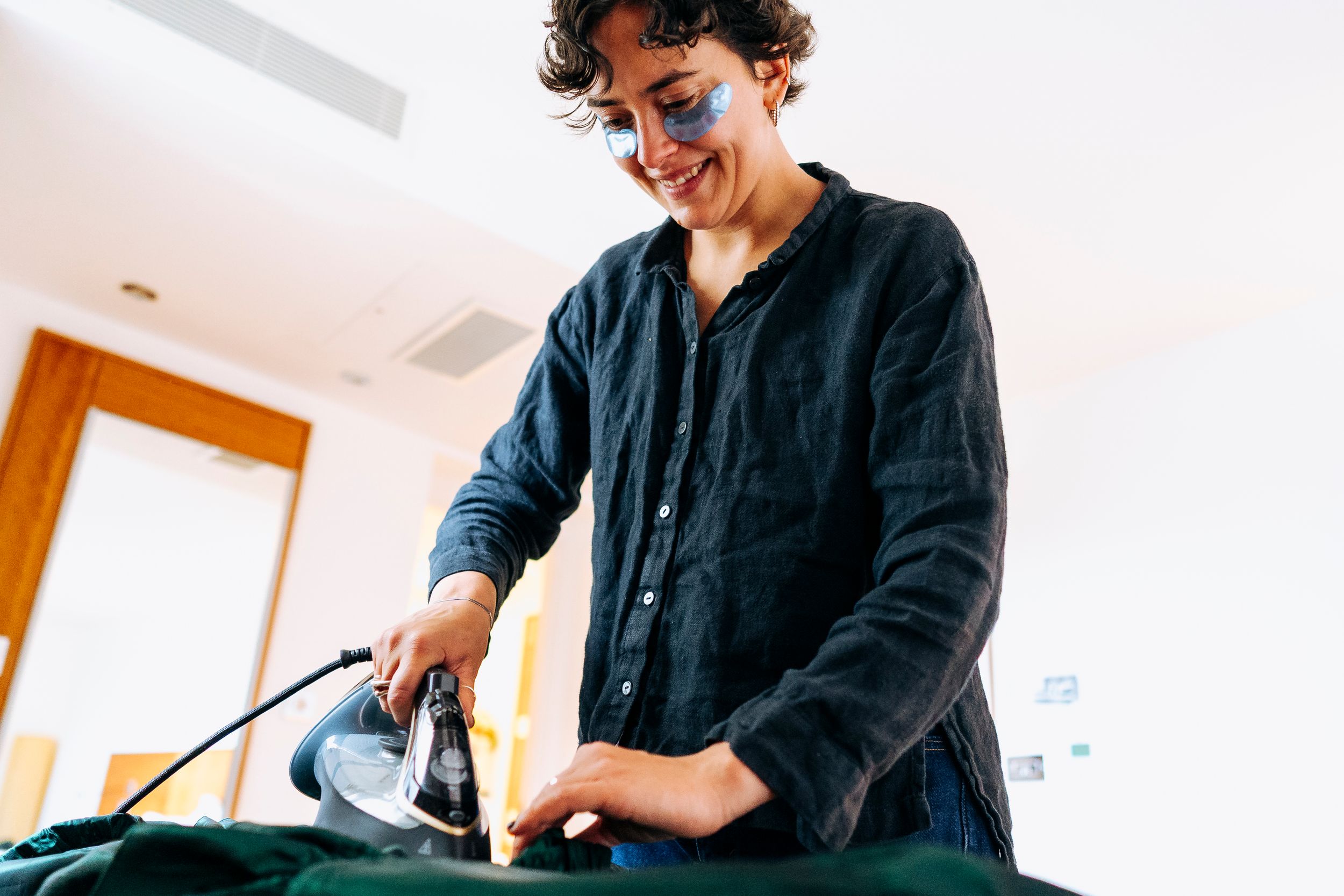 Smiling person with curly hair ironing green fabric garment in bright room before wedding.