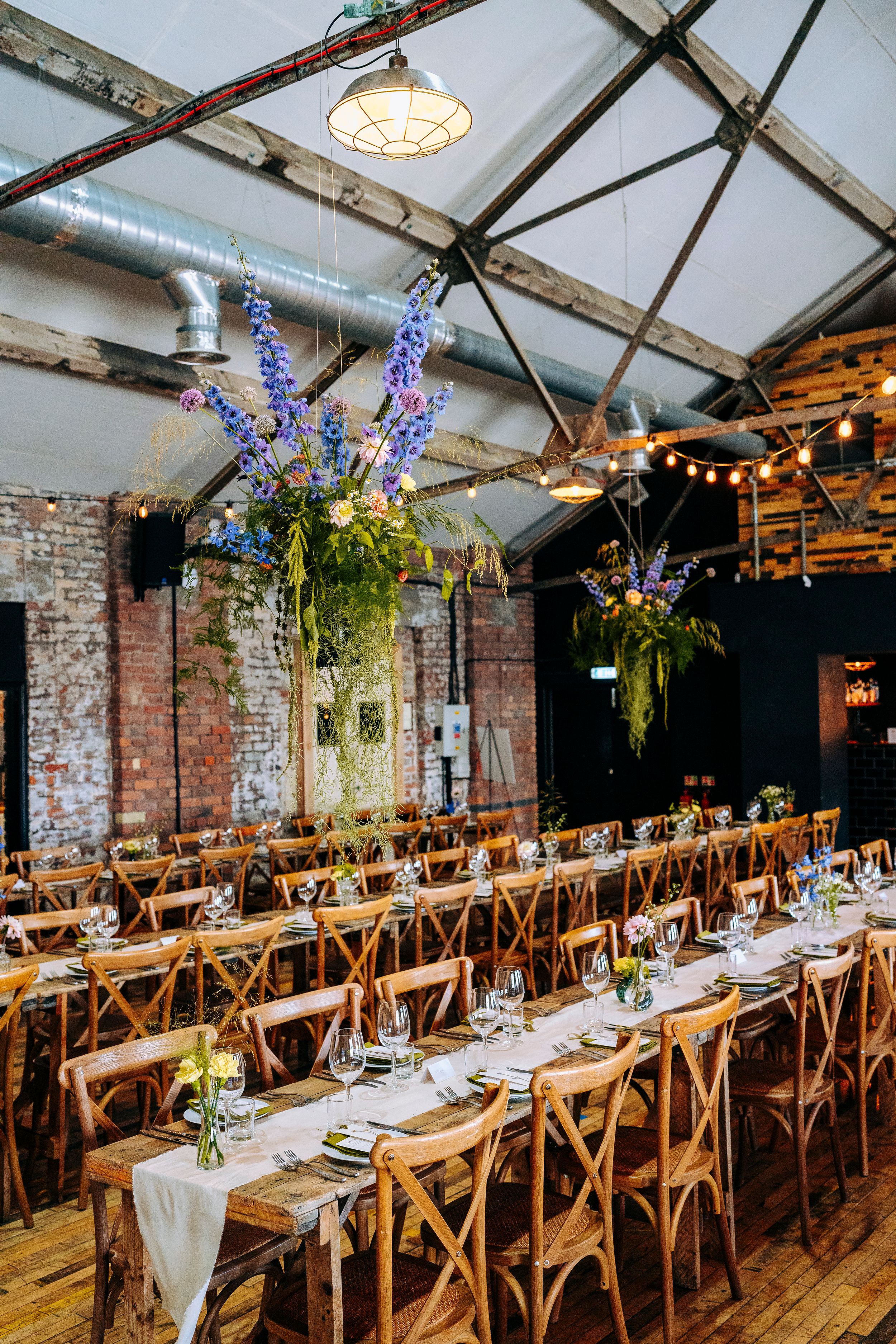 Rustic wedding reception venue with long wooden tables, cross-back chairs, blue floral arrangements, exposed brick walls, and string lights.