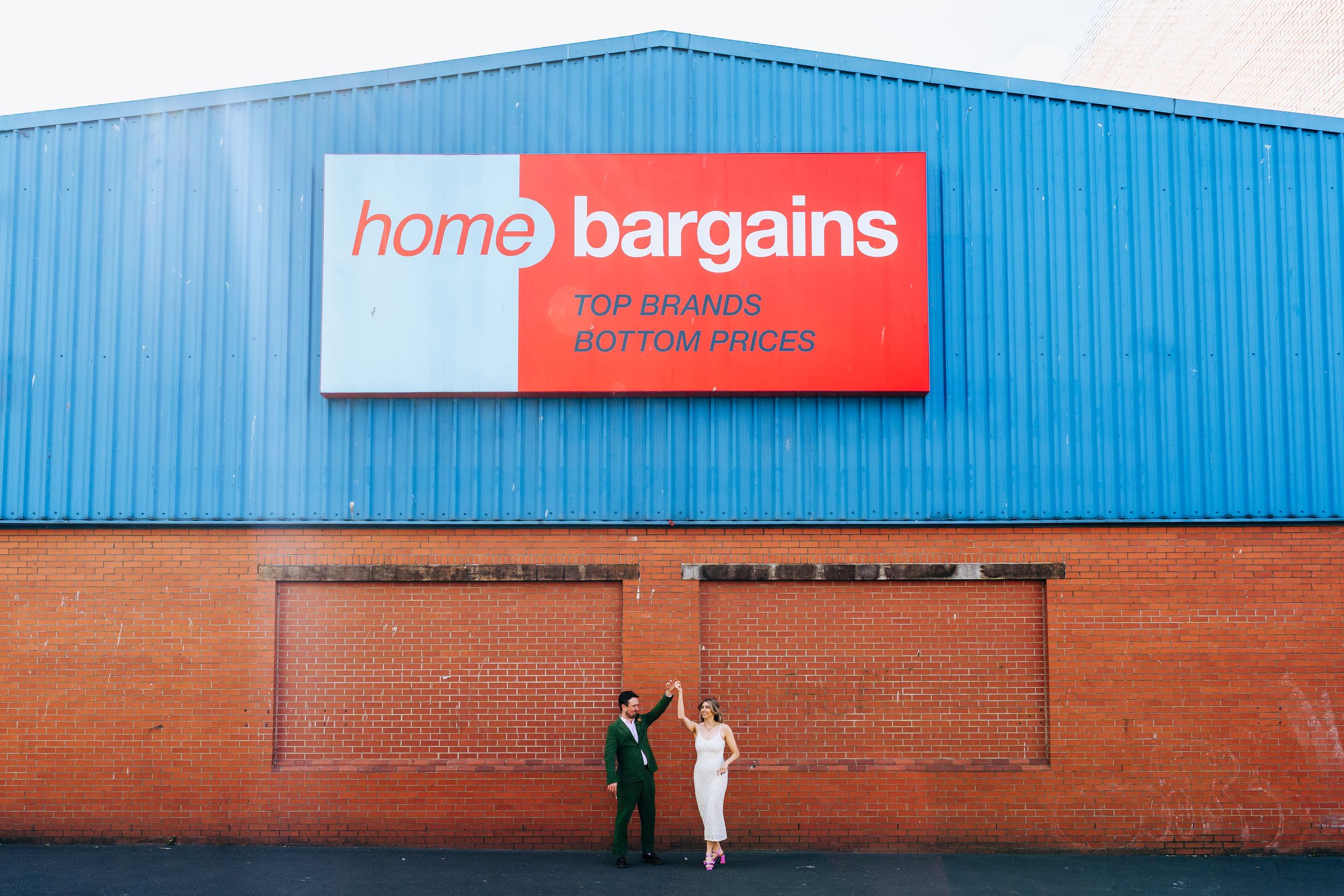 Bride and groom pose playfully in front of a large Home Bargains store sign outdoors in Liverpool.