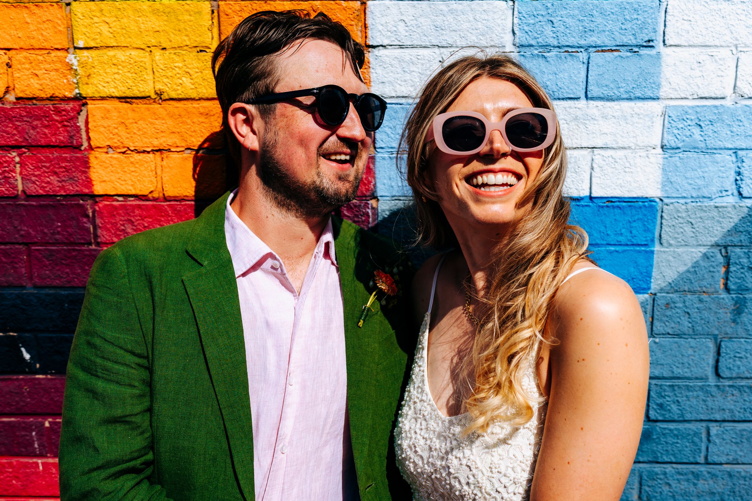 Smiling couple wearing sunglasses posing together in front of a vibrant colorful painted brick wall outdoors.