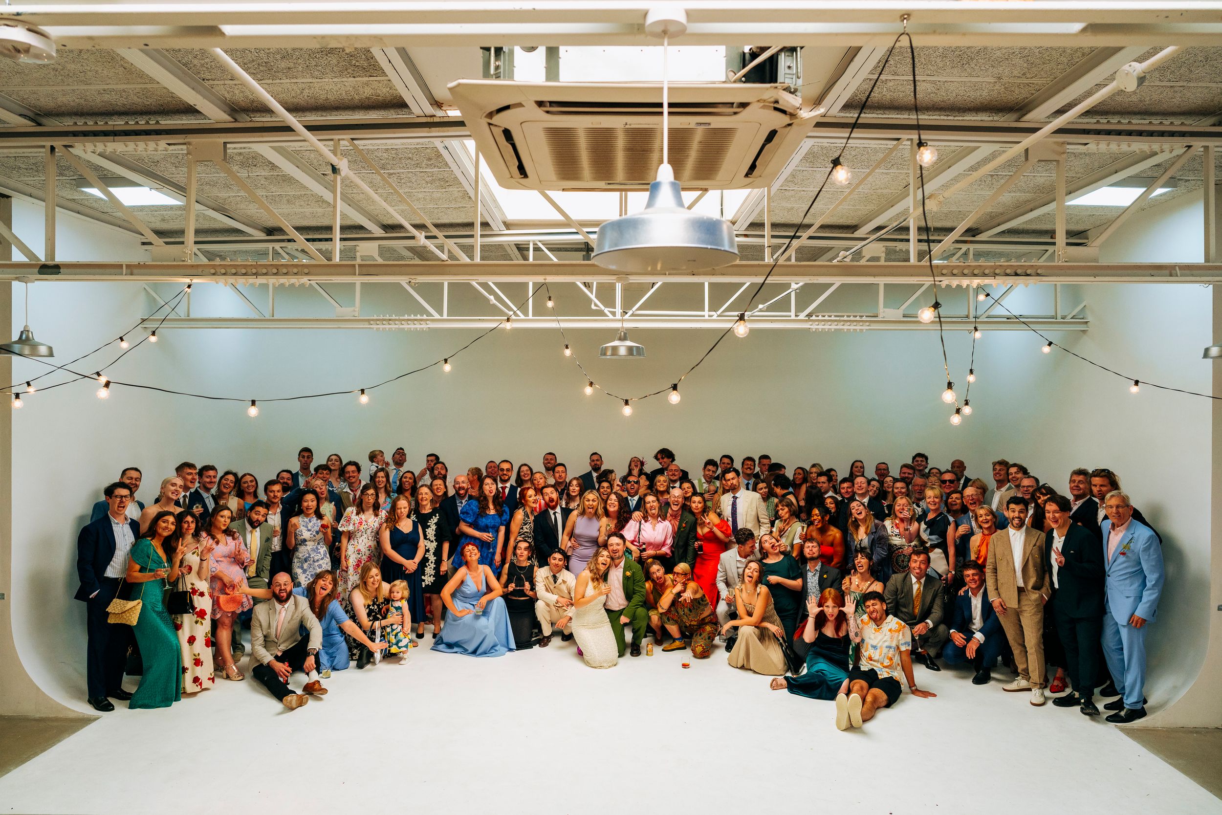 Large wedding group photo with all guests gathered together in a bright white studio venue with string lights.