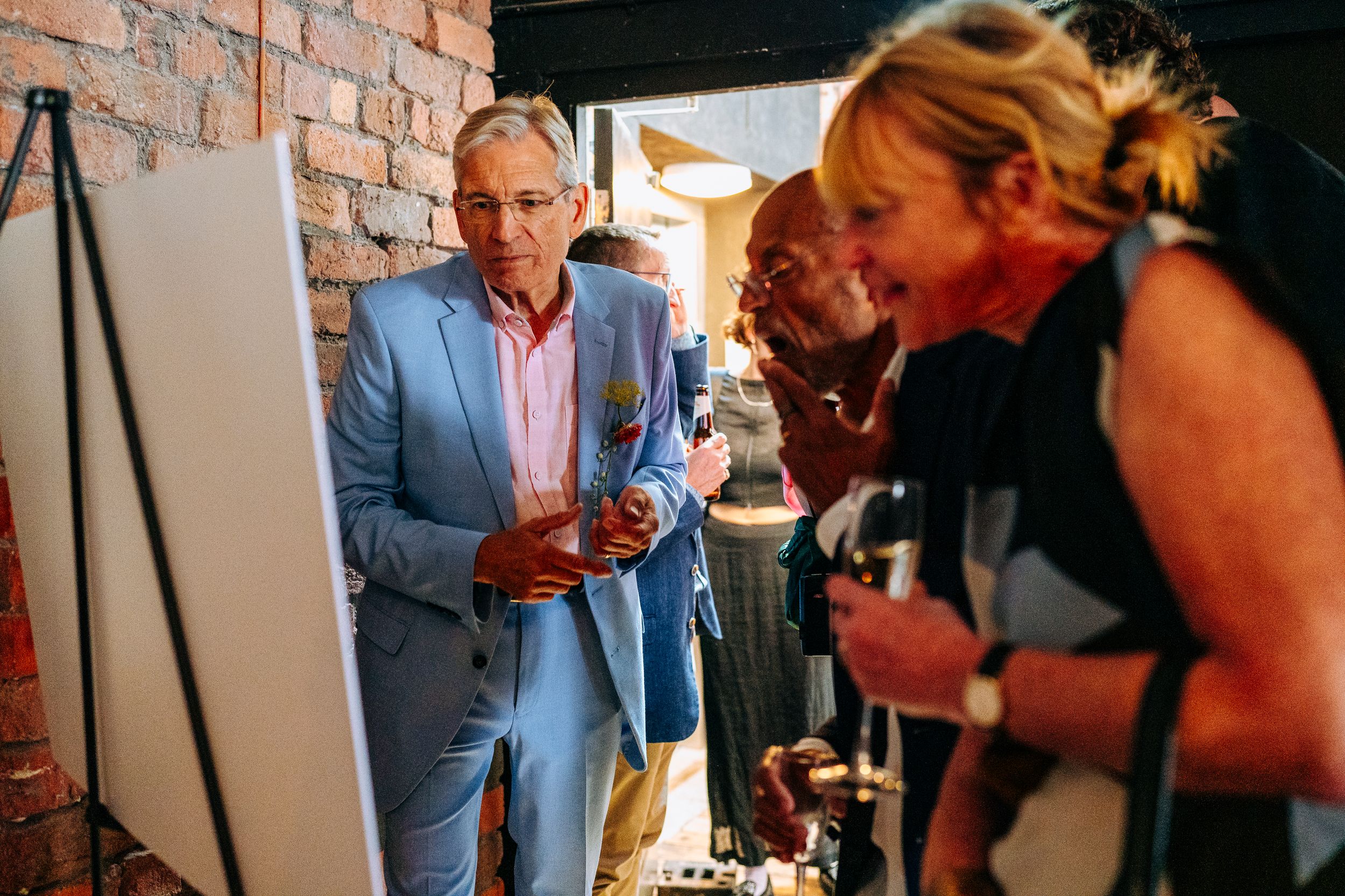 Wedding guest in light blue suit examining a display board at a rustic brick-walled wedding reception venue.