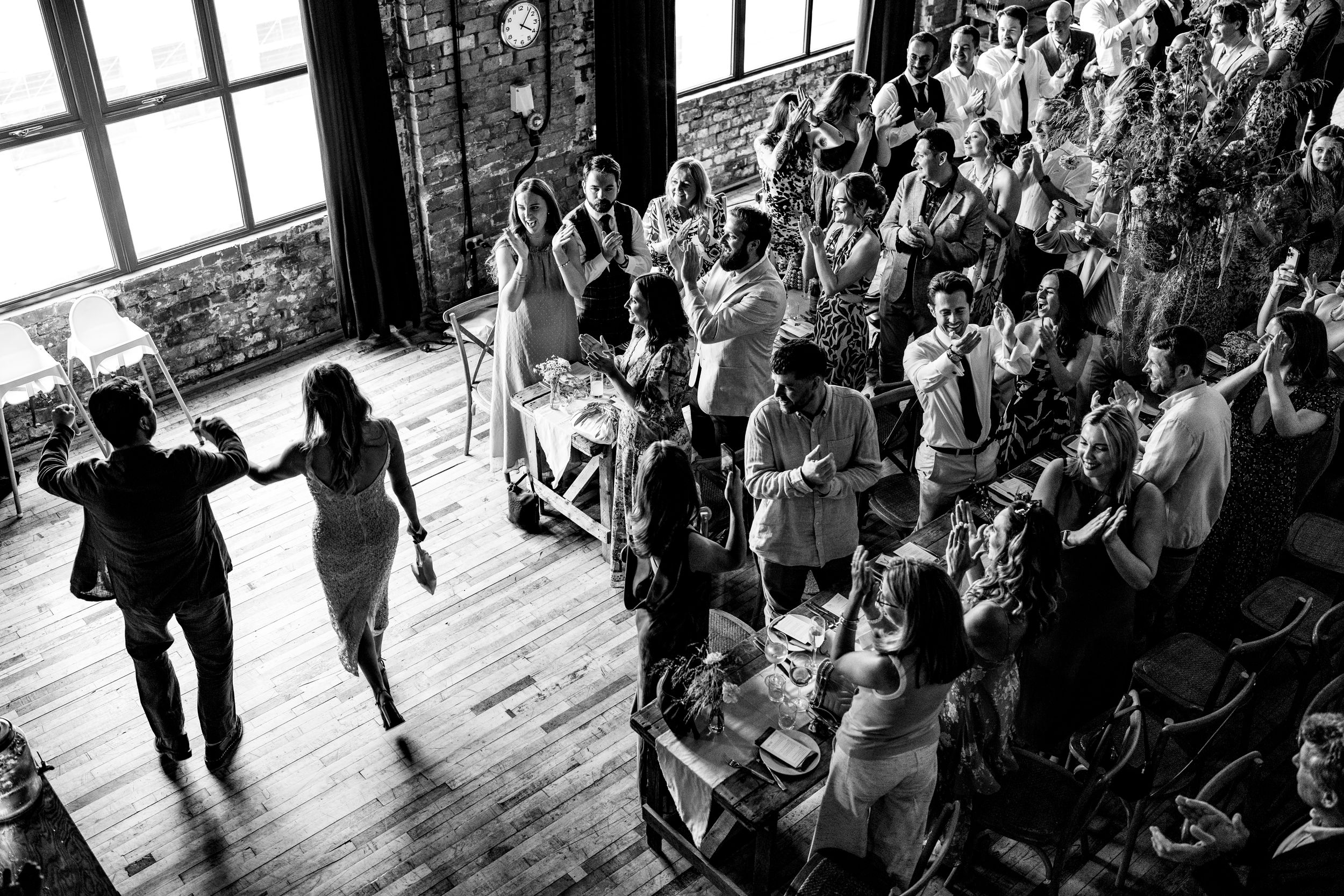 Black and white aerial view of bride and groom entering wedding reception as guests cheer in industrial venue.