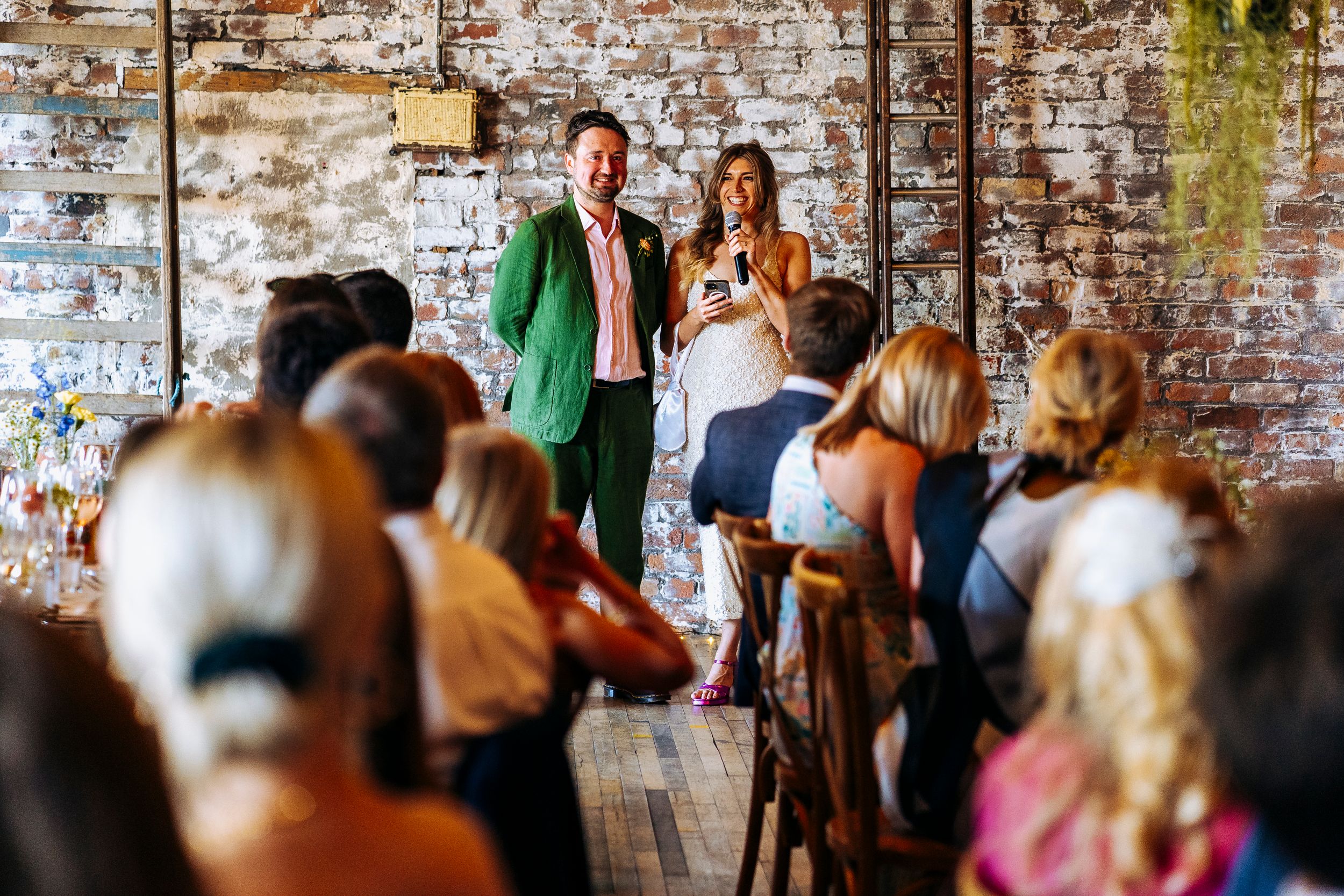 Groom in green suit and bride giving speeches to seated wedding guests in a rustic brick-walled reception venue.