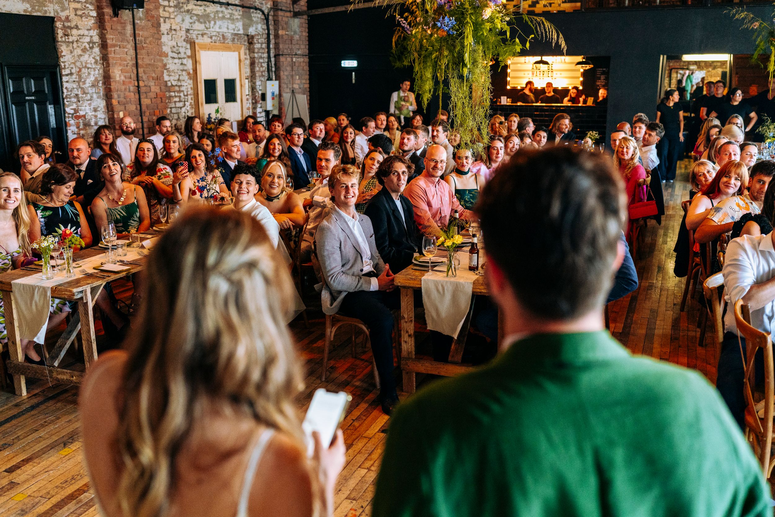 Bride and groom addressing a large crowd of seated wedding guests in a warmly lit rustic reception hall.
