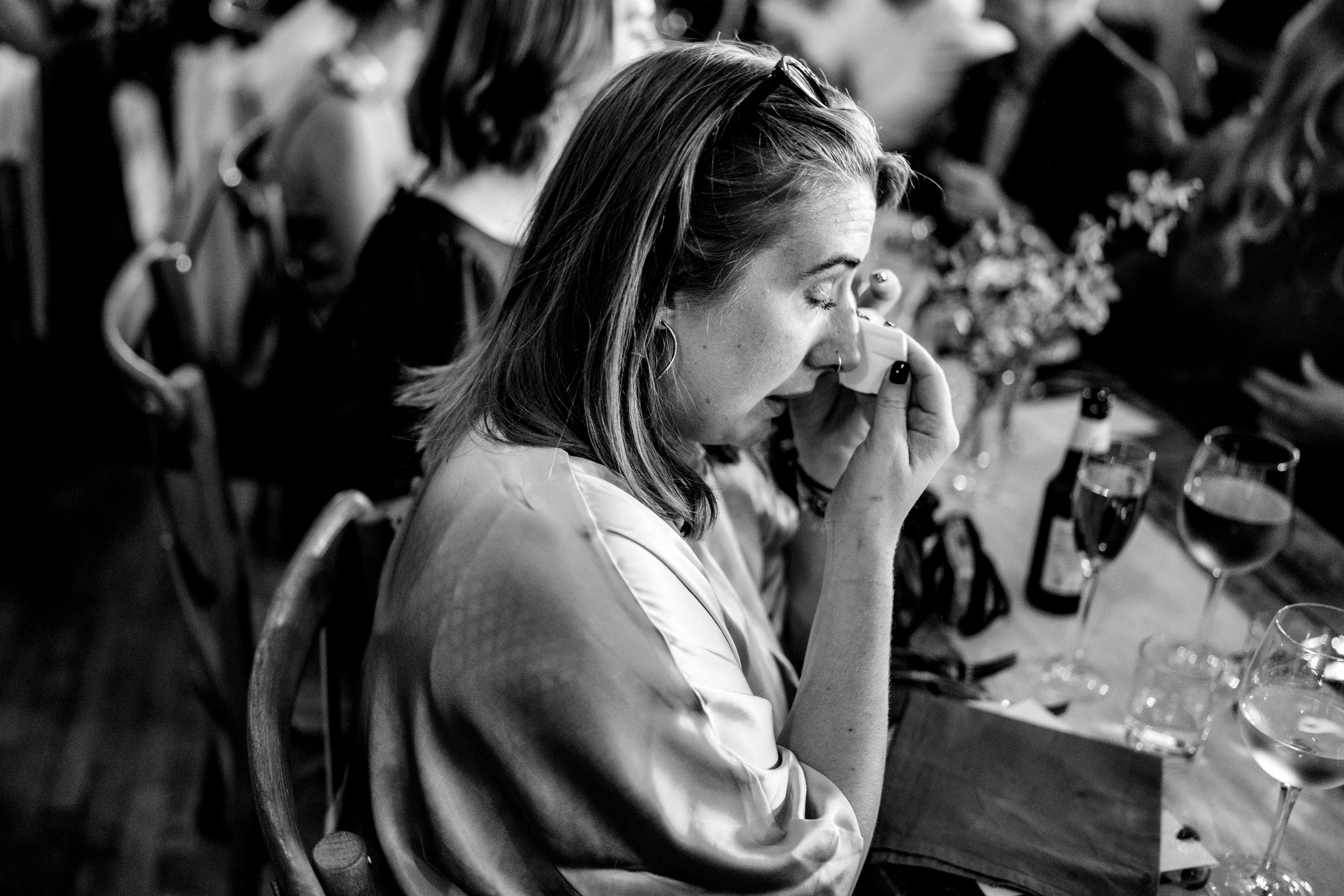 Emotional wedding guest wiping tears at reception table in candid black and white photography moment.