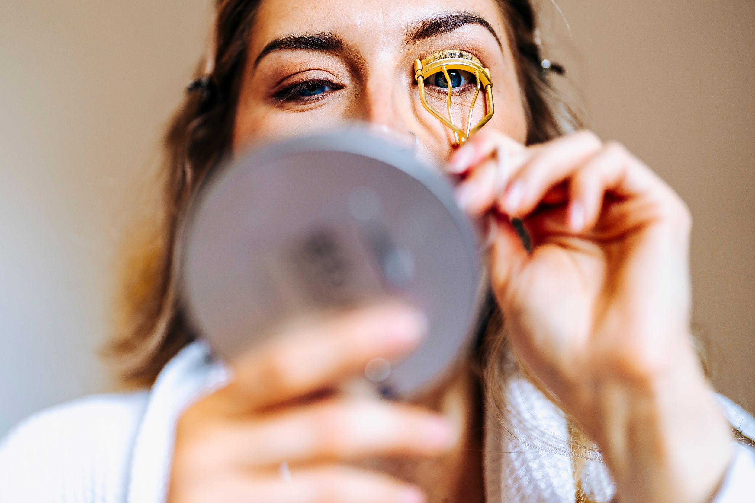 A woman curls her eyelashes using a gold eyelash curler while looking into a small mirror.