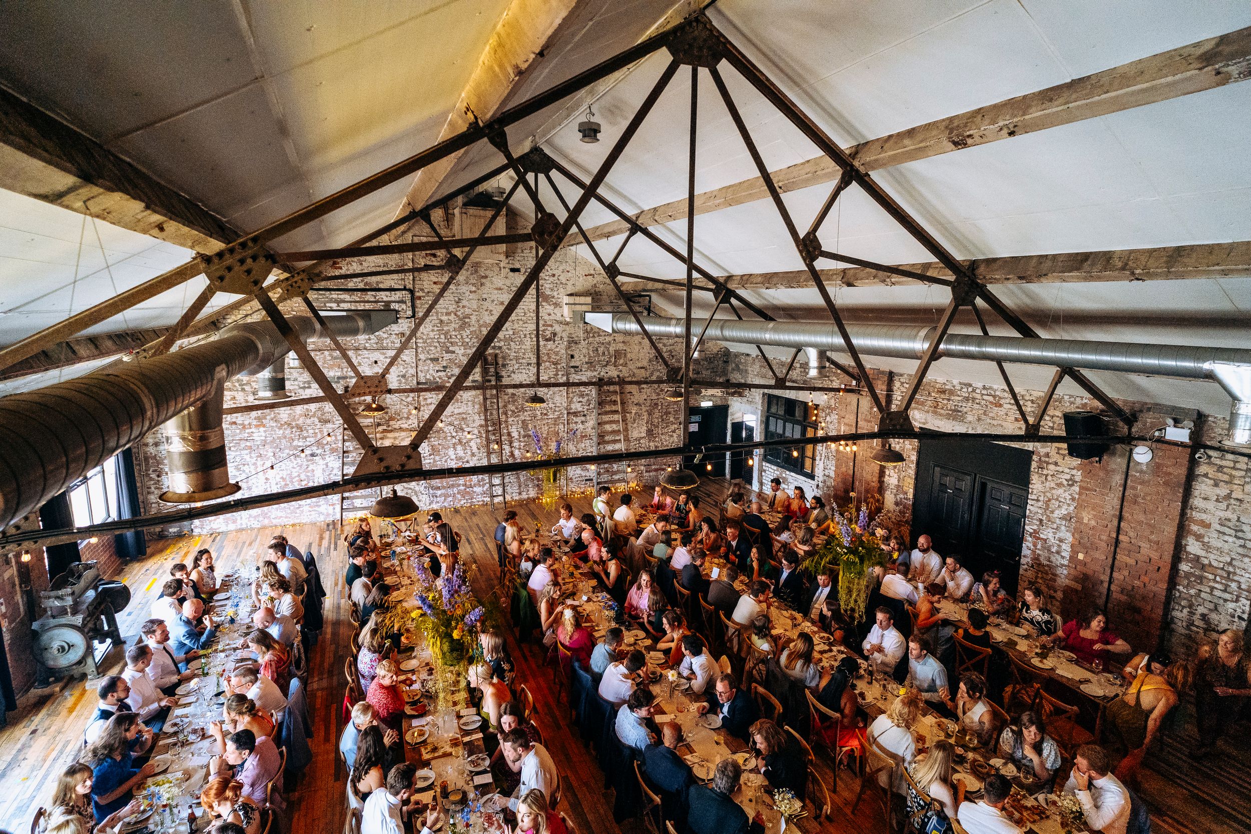 Aerial view of packed wedding reception with long banquet tables full of guests in a rustic industrial venue.