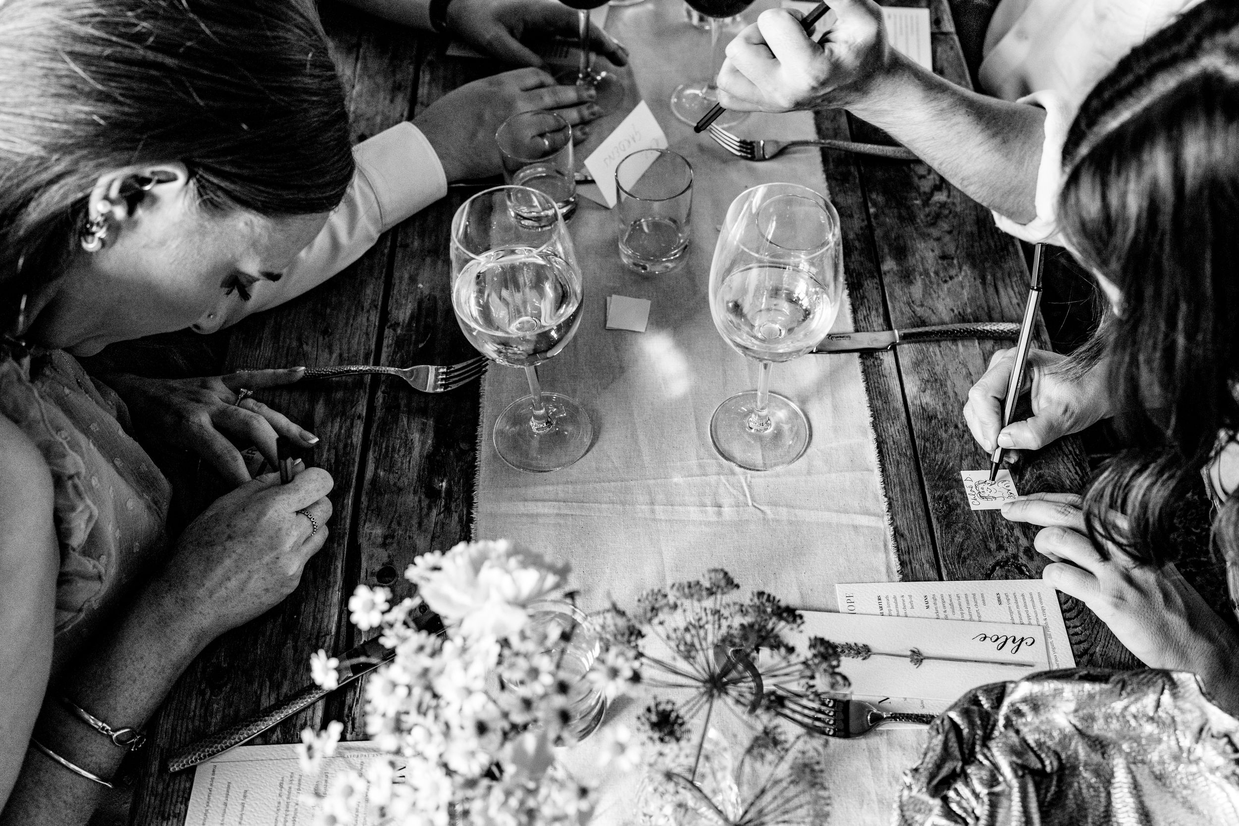 Black and white overhead shot of wedding guests writing notes beside wine glasses on a rustic wooden table.