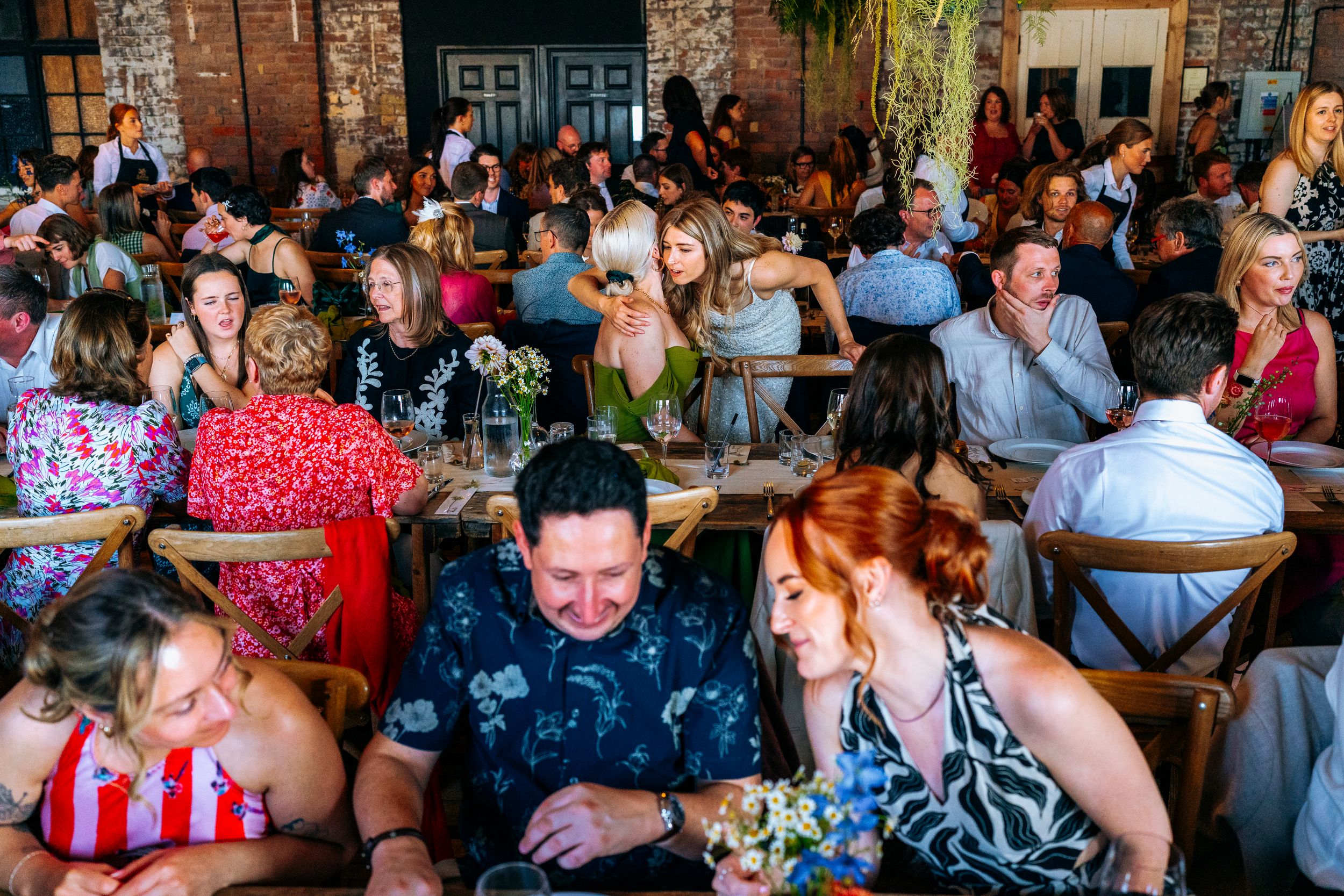 Large crowd of wedding guests socializing and laughing at decorated reception tables indoors.