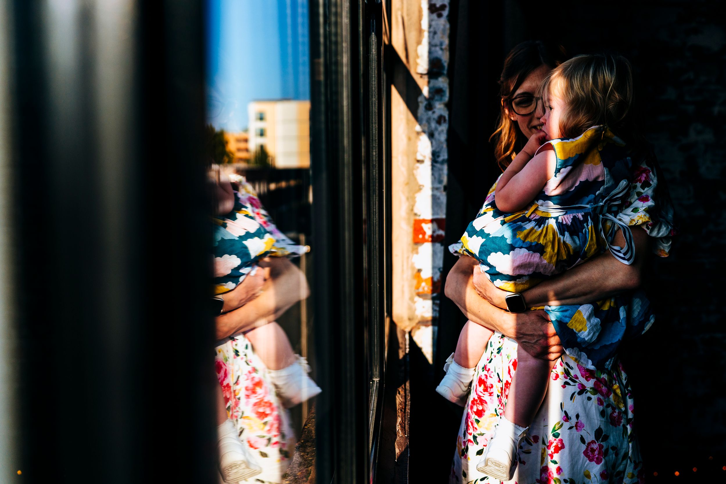 Woman in floral dress holding a baby reflected in a sunlit window at golden hour.