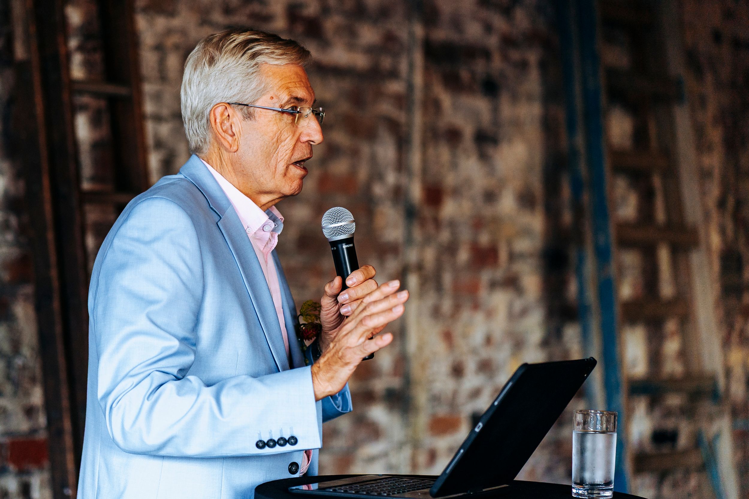 Older man in light blue blazer speaking into a microphone at an indoor event podium.