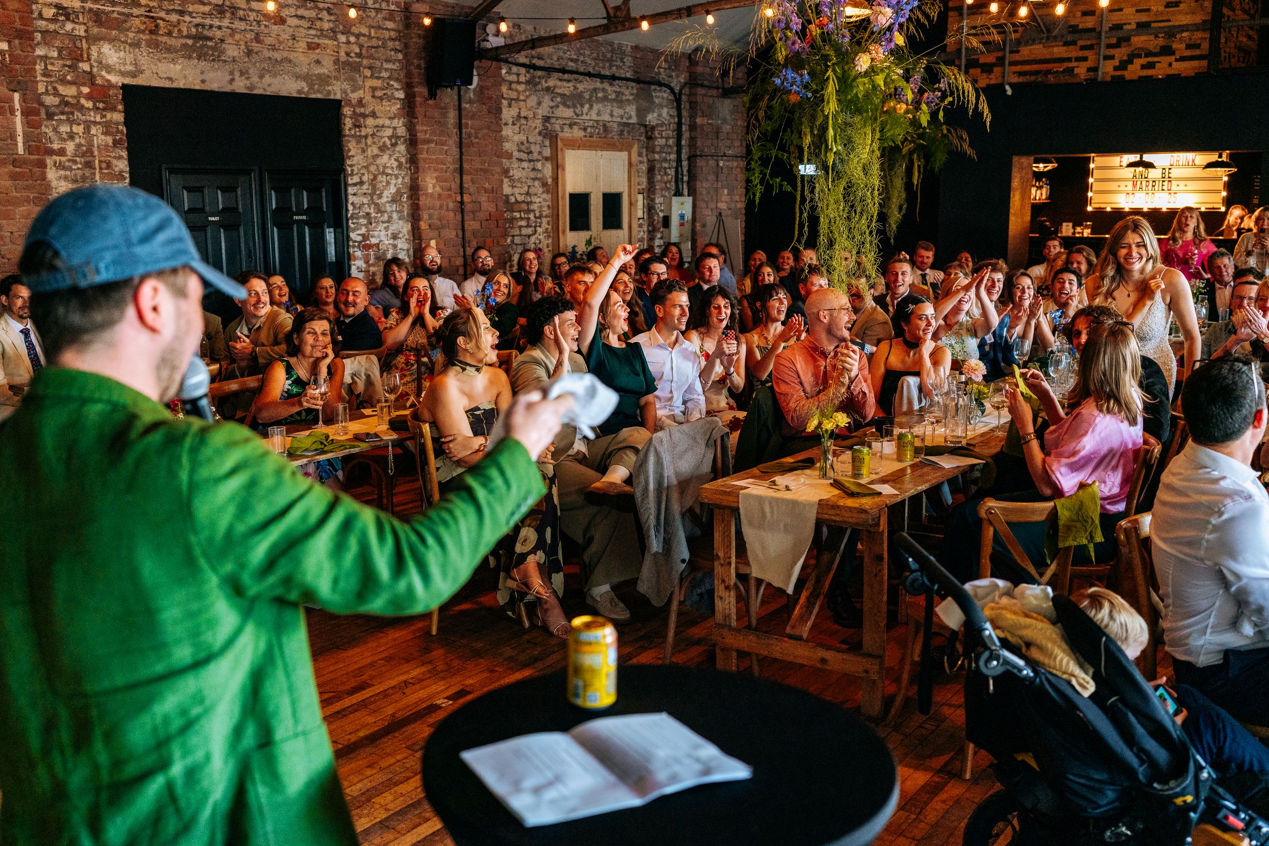 Wedding MC in green jacket addresses applauding guests at elegant rustic brick venue reception.