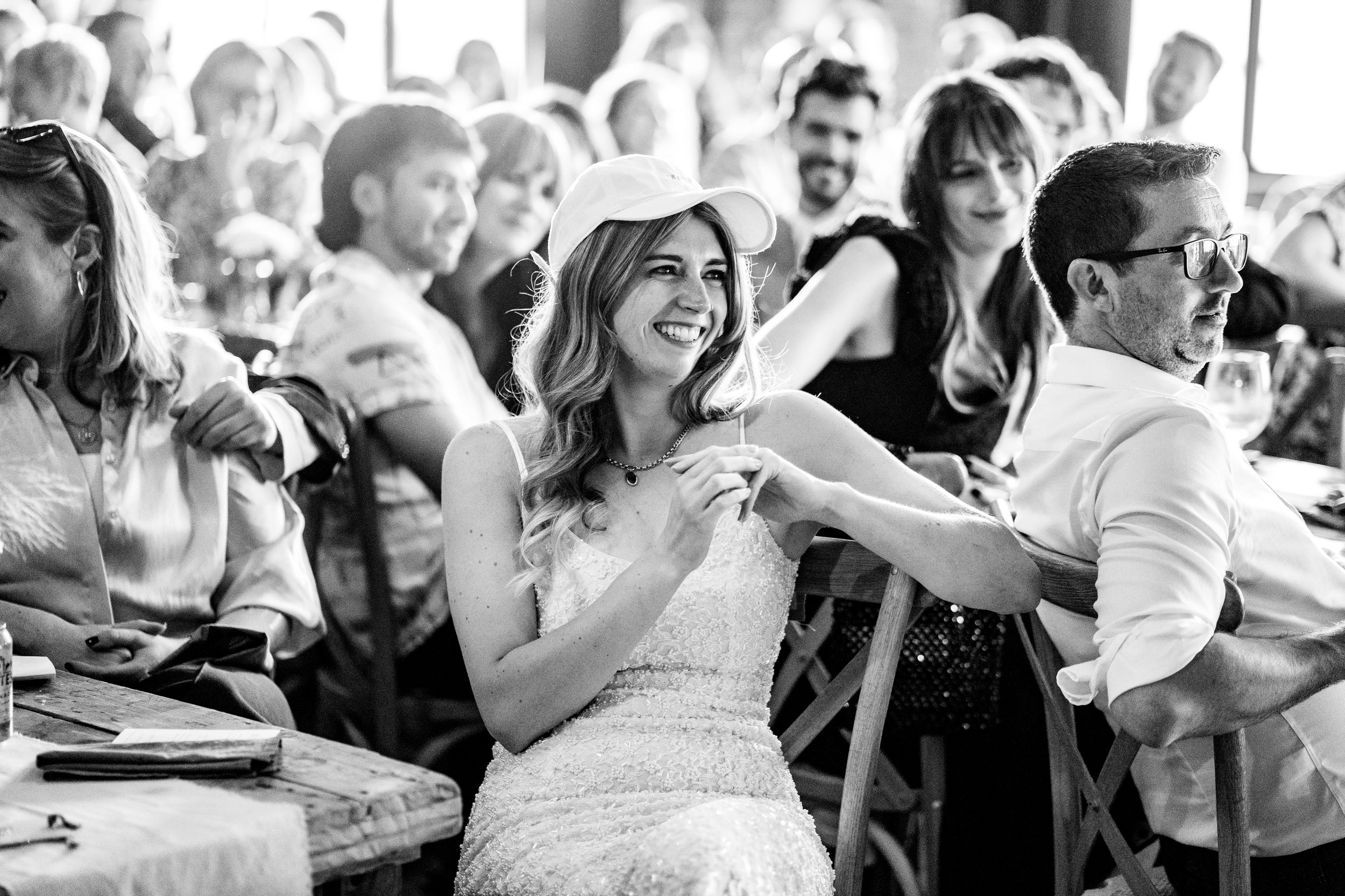 Black and white image of smiling bride in lace dress seated with guests at reception.