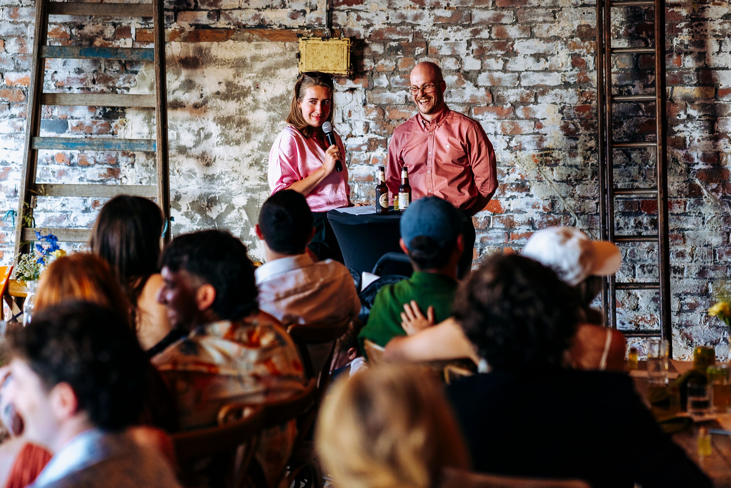 Couple giving speech at wedding reception in rustic brick venue with seated guests listening attentively.