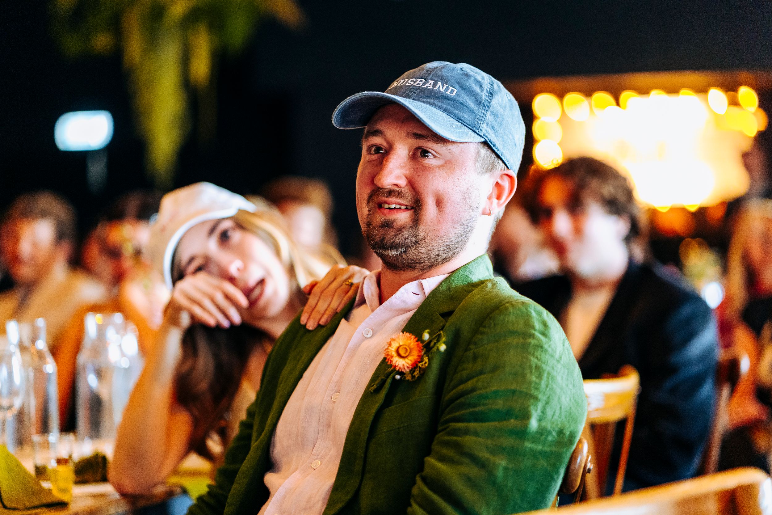 Smiling wedding guest in green blazer and blue cap relaxing at decorated reception table.