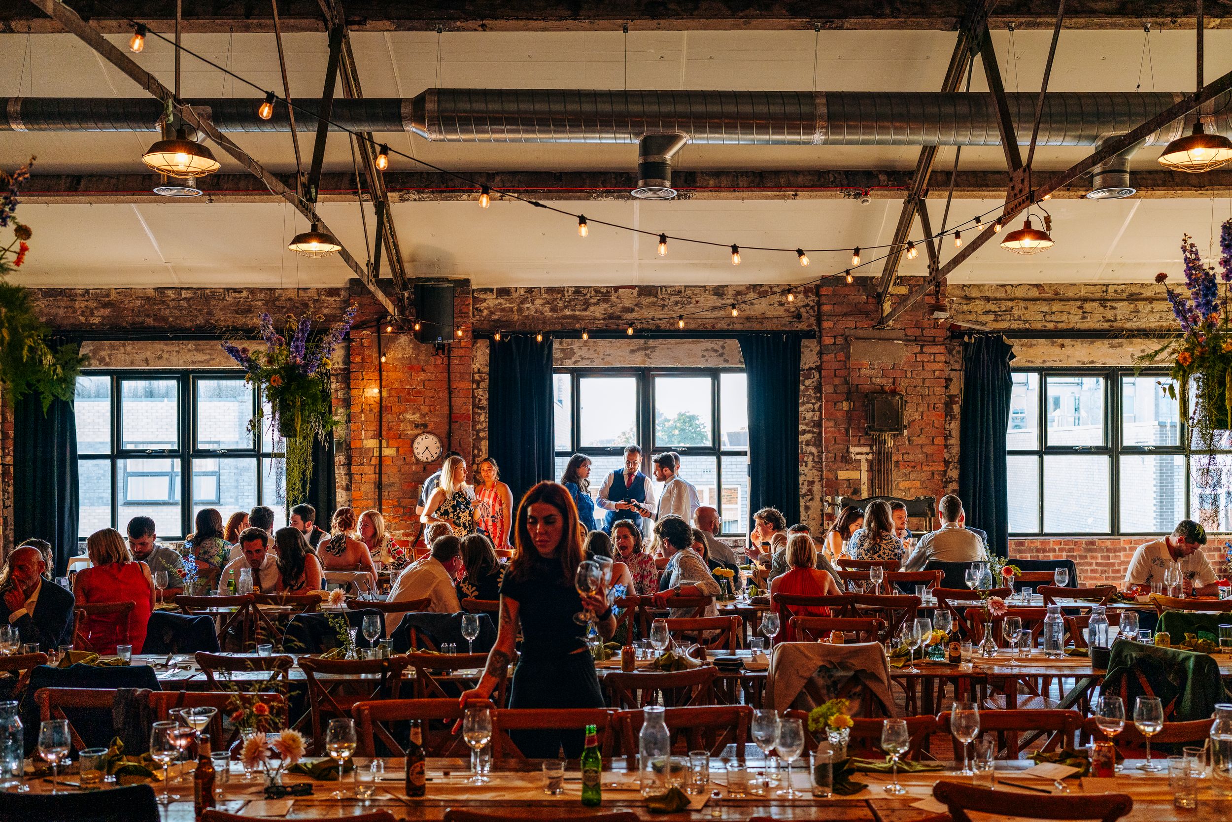 Guests dining at long tables in an industrial brick wedding reception venue with string lights.