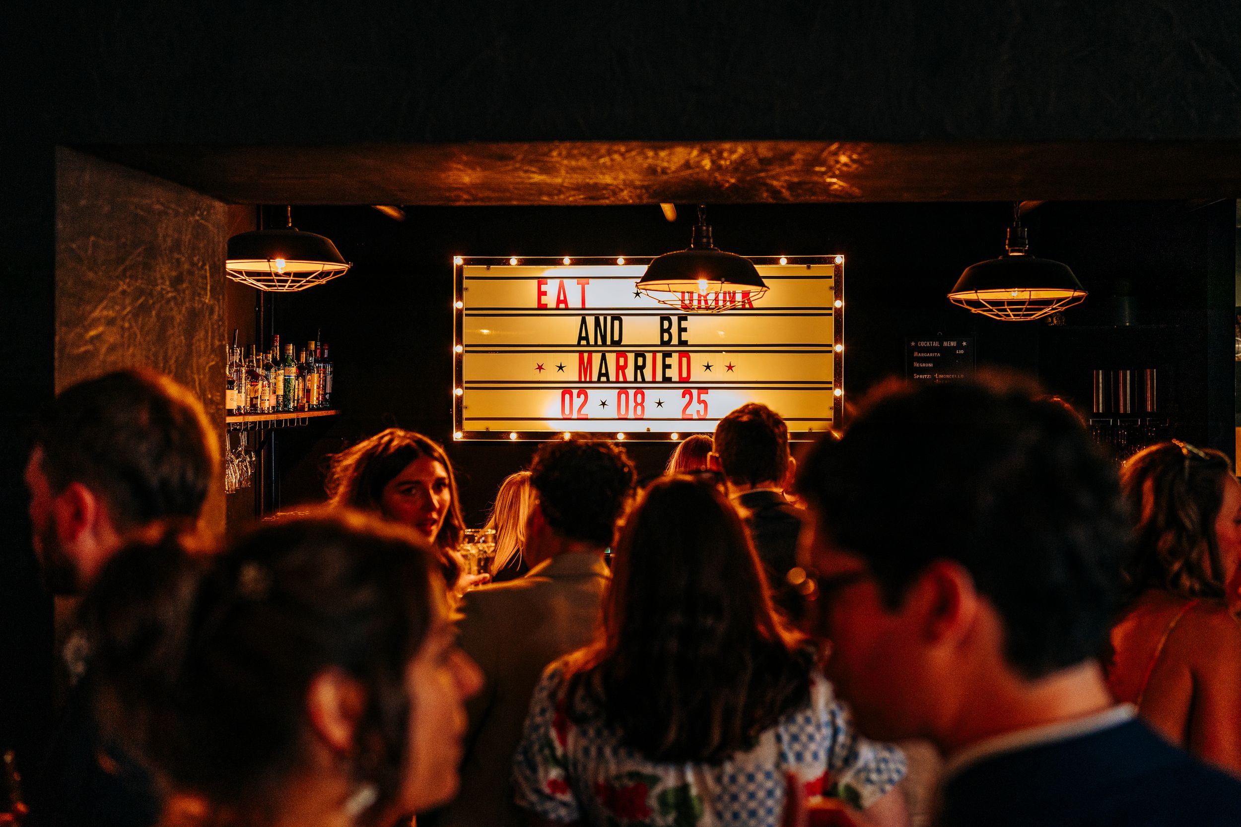 Wedding guests gathered around illuminated marquee sign reading just married at evening reception bar.