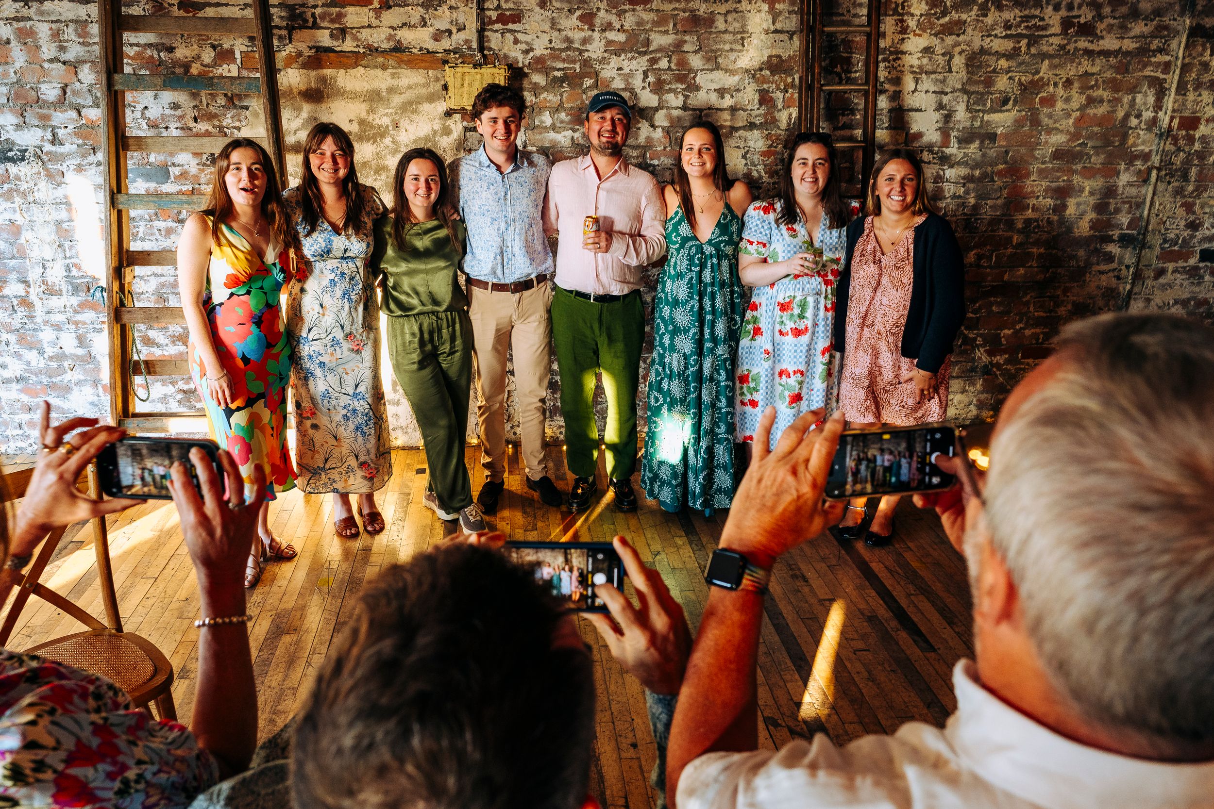 Group of friends posing for photos at a rustic brick wedding venue while guests photograph them.