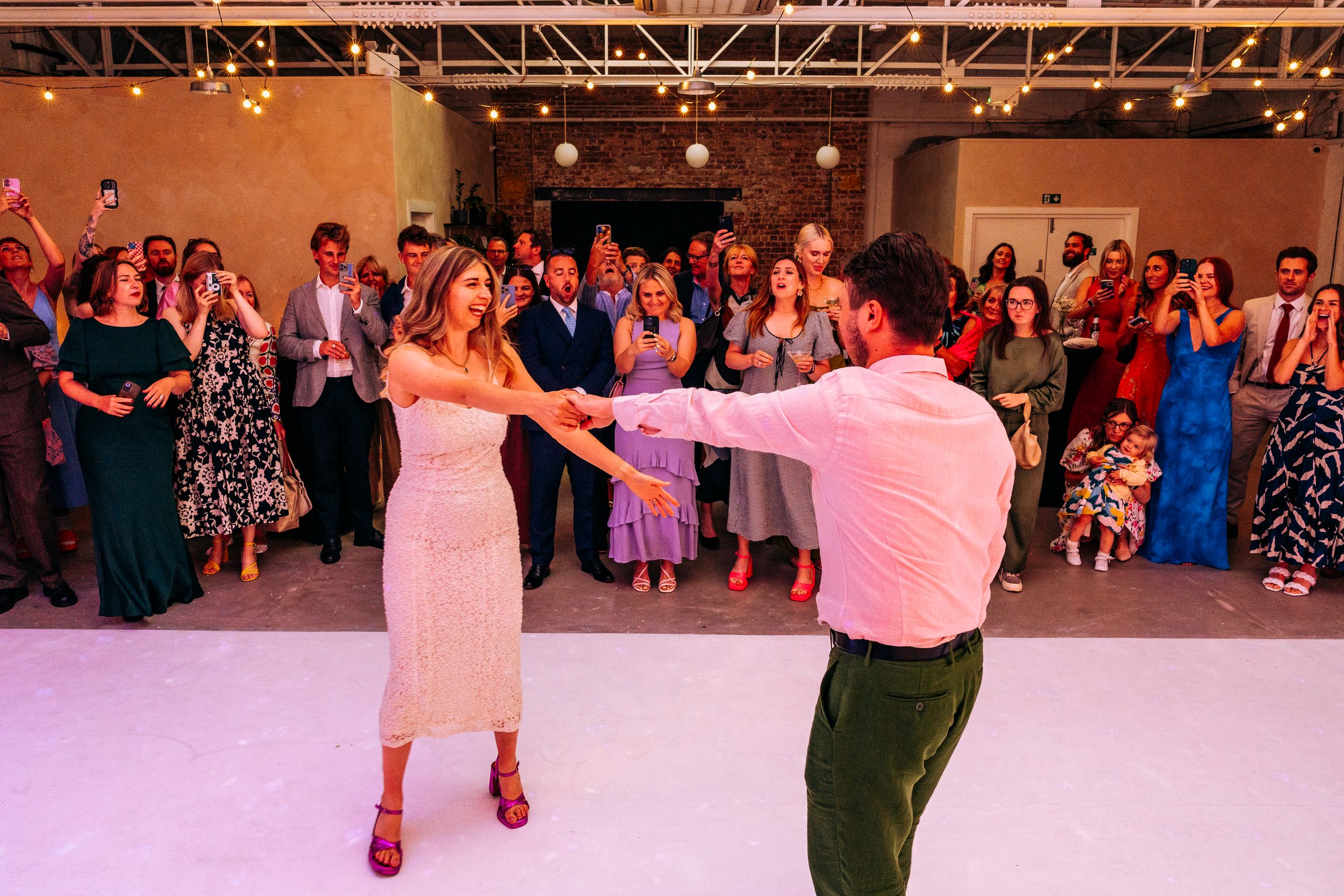 Bride and groom share their first dance while wedding guests cheer in a bright venue.