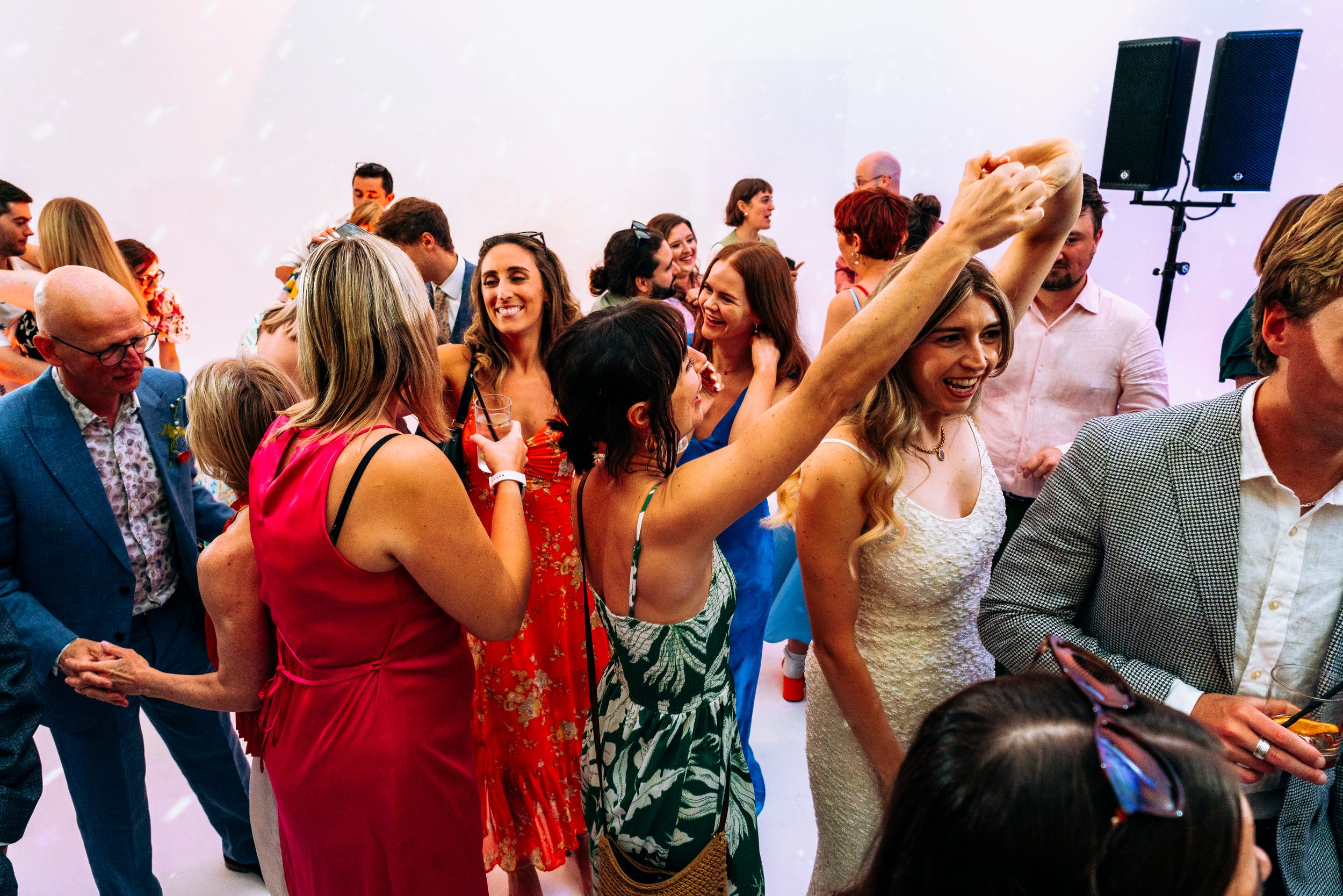 Bride in lace dress dancing joyfully surrounded by colorfully dressed wedding reception guests.