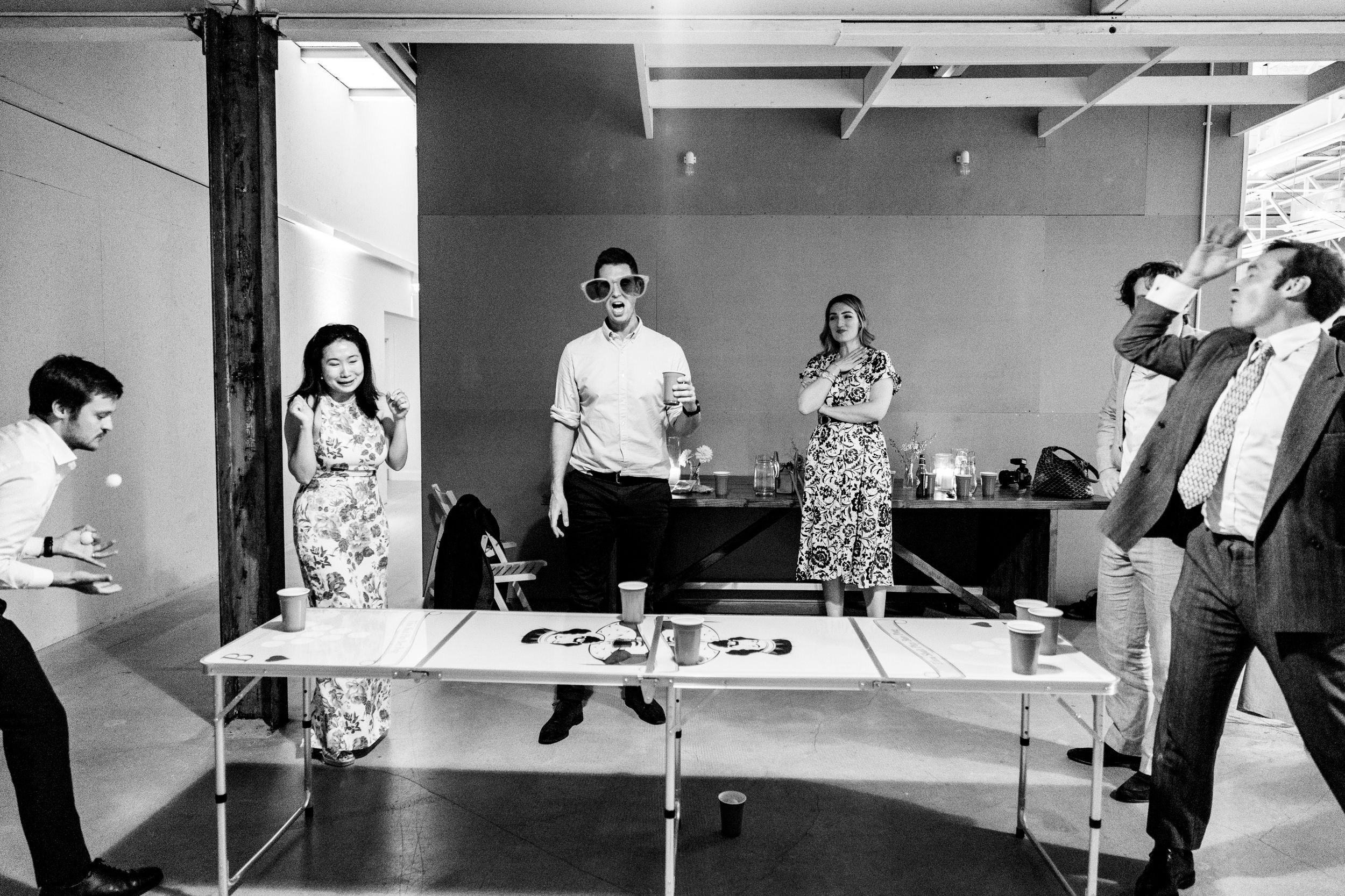 Guests play beer pong at an indoor wedding reception in a black and white photo.