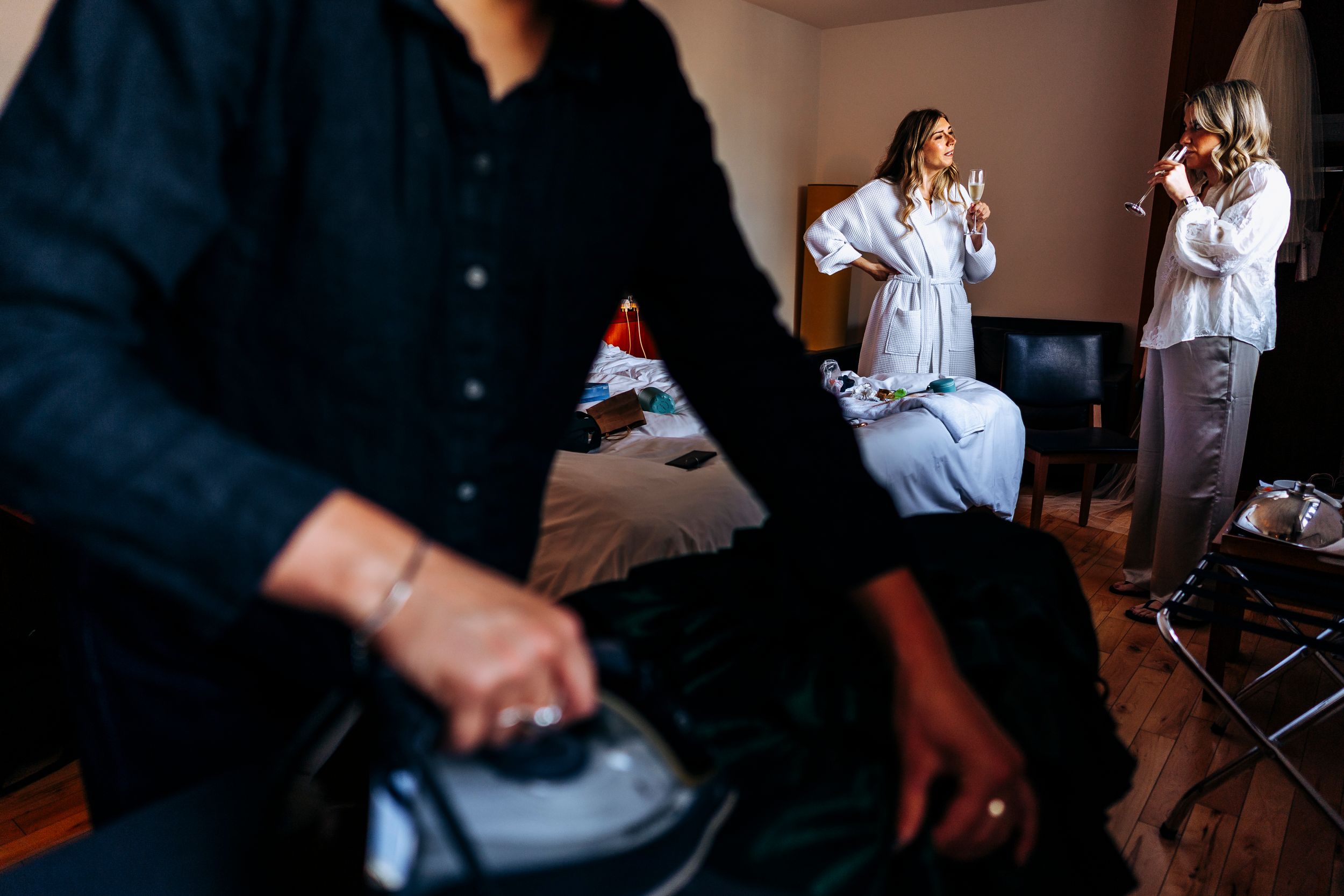 A bride in a white robe stands in a hotel room while preparations are made around her.