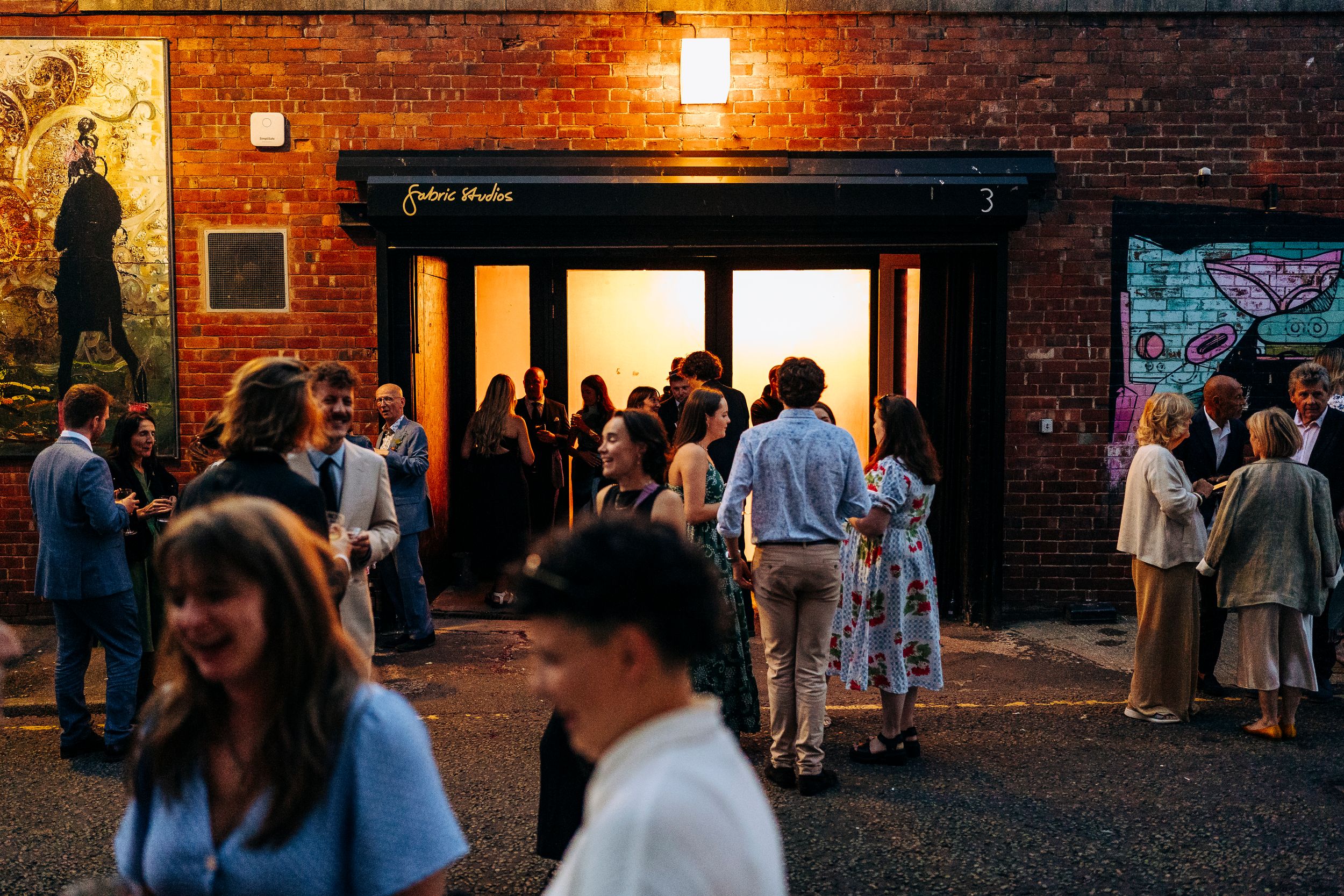 Guests mingle outside a brick venue with colorful murals at an evening wedding reception.