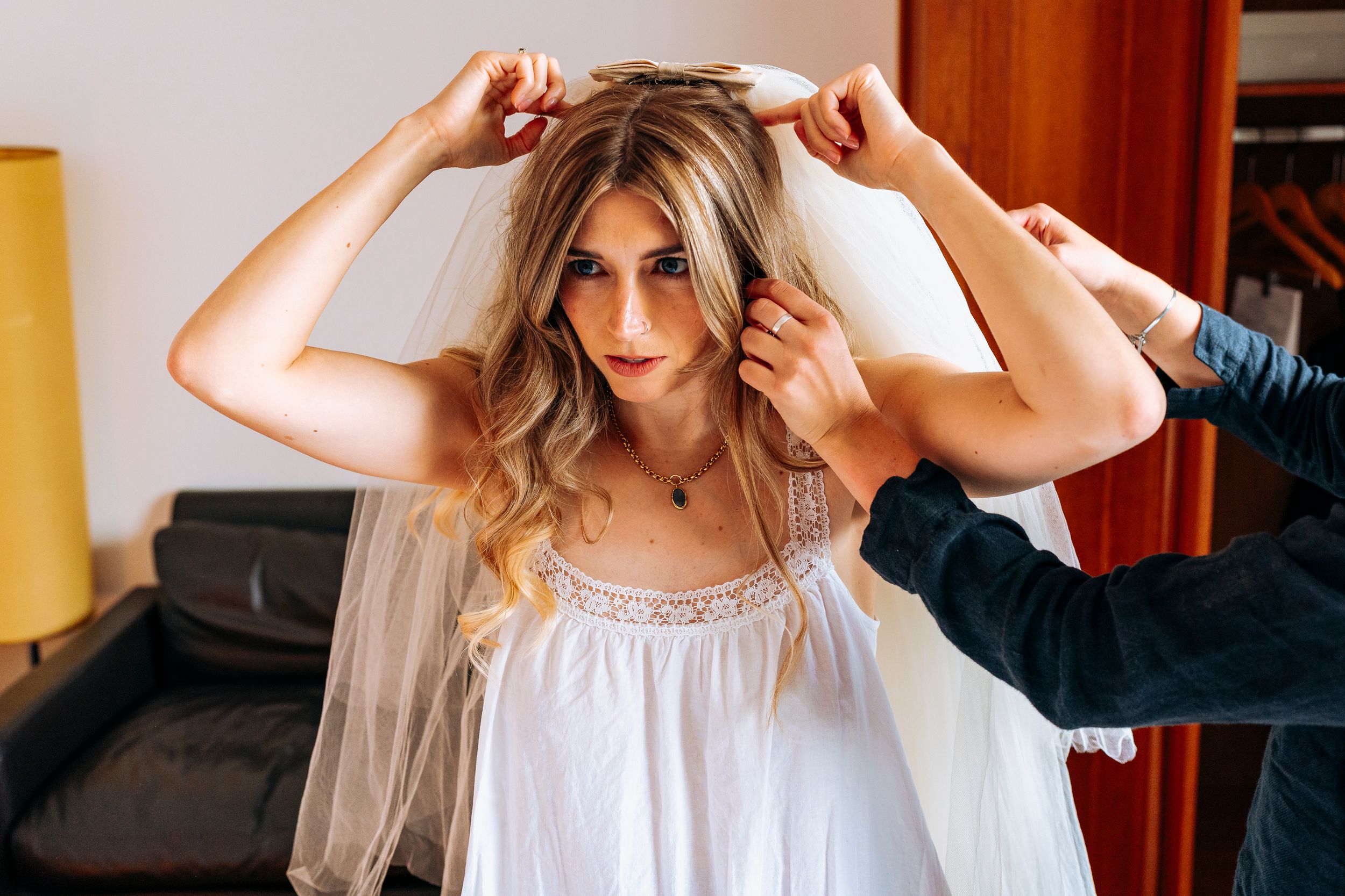A blonde bride has her wedding veil placed on her head by a stylist indoors.