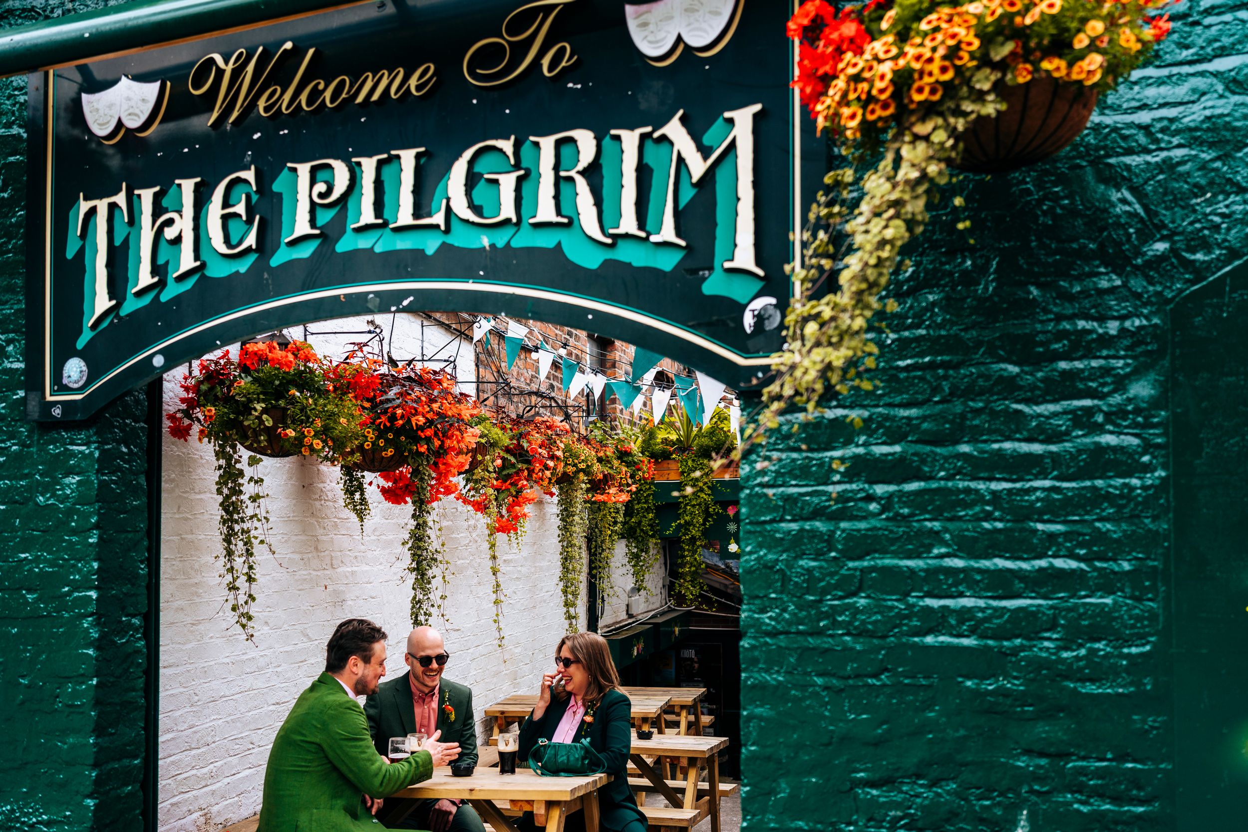 Three guests sit beneath a floral-adorned entrance to The Pilgrim pub during a wedding celebration.