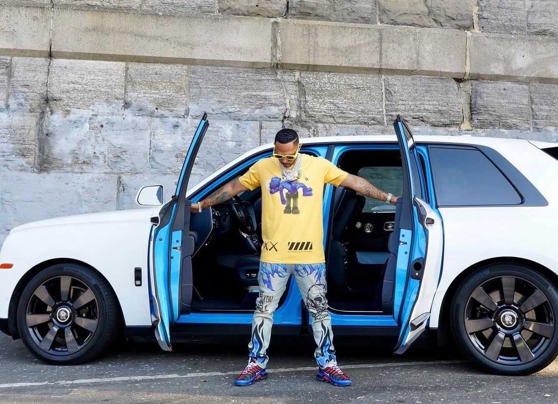 Man in yellow jersey posing beside white and blue Rolls Royce Cullinan SUV with doors open.