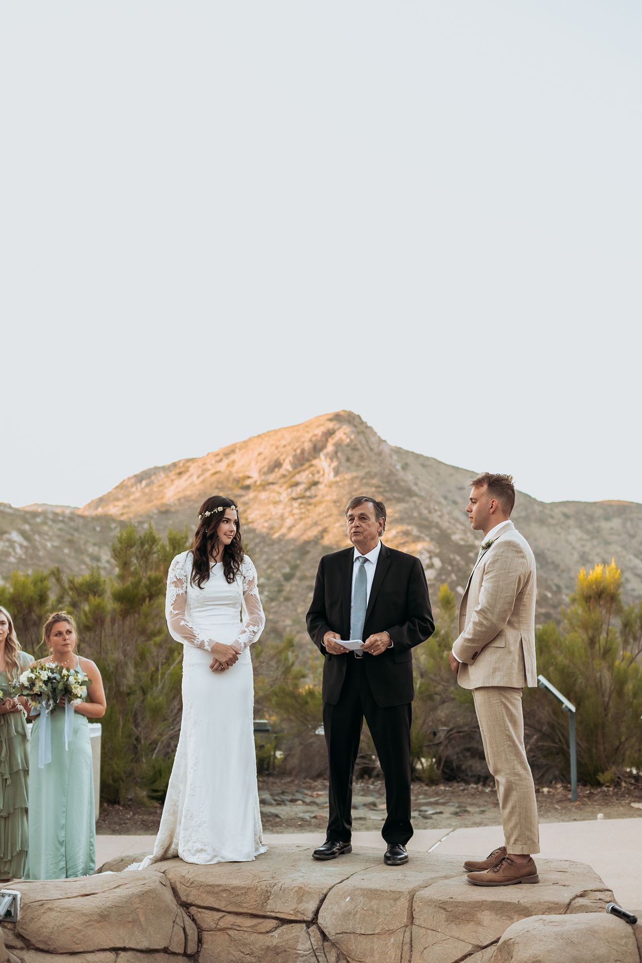 Outdoor wedding ceremony on rocks with bride in white lace gown, groom in tan suit, and officiant, mountain backdrop.