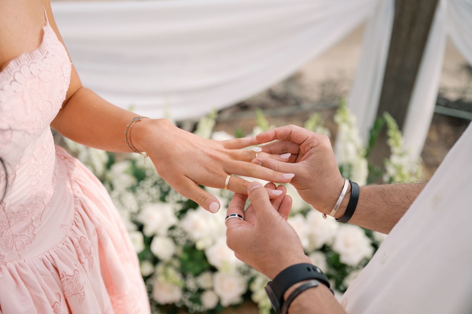 Groom placing a wedding ring on bride's finger during outdoor ceremony with white floral backdrop.