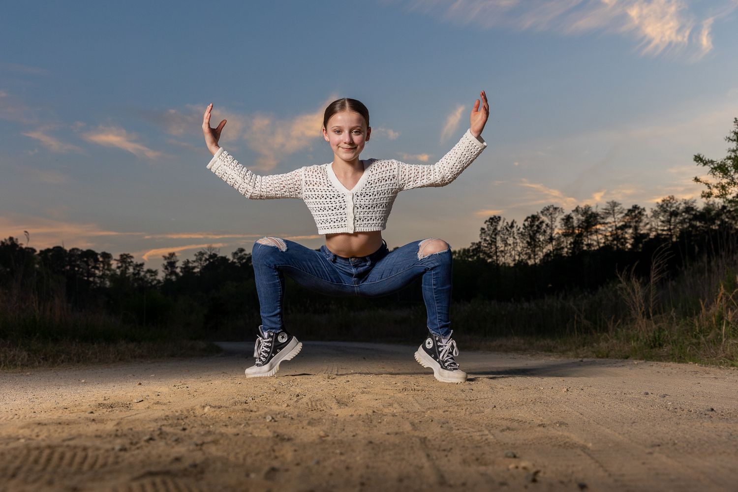 Young girl in white crop top and jeans doing a squat pose on sandy ground at dusk with trees in background.