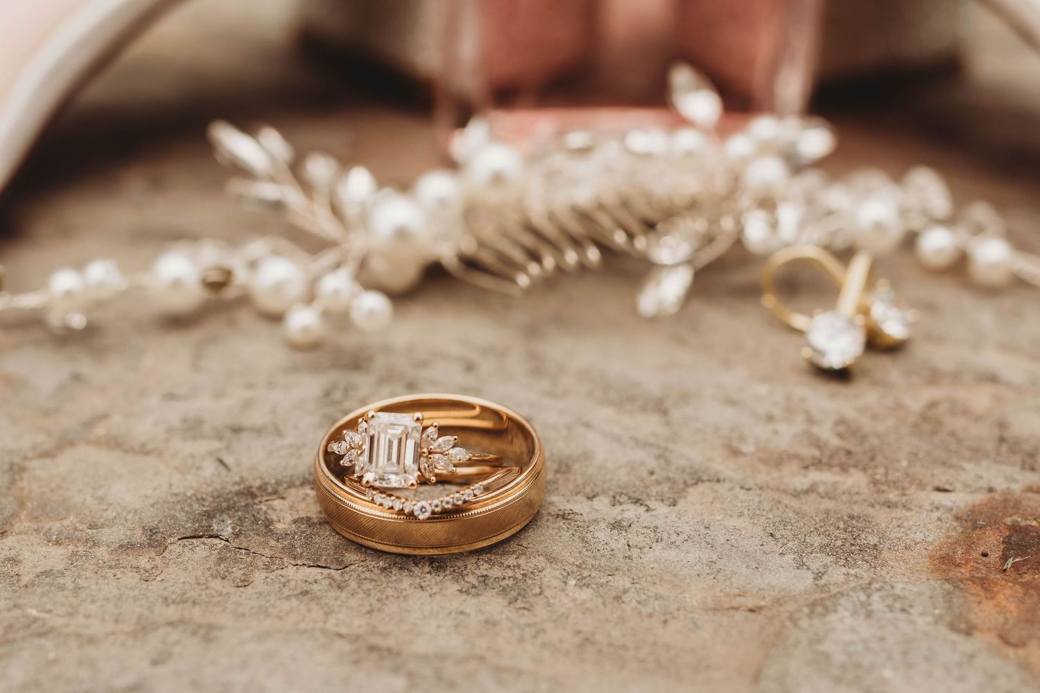 Gold wedding rings and delicate floral hair accessories rest on a textured beige surface in warm lighting.
