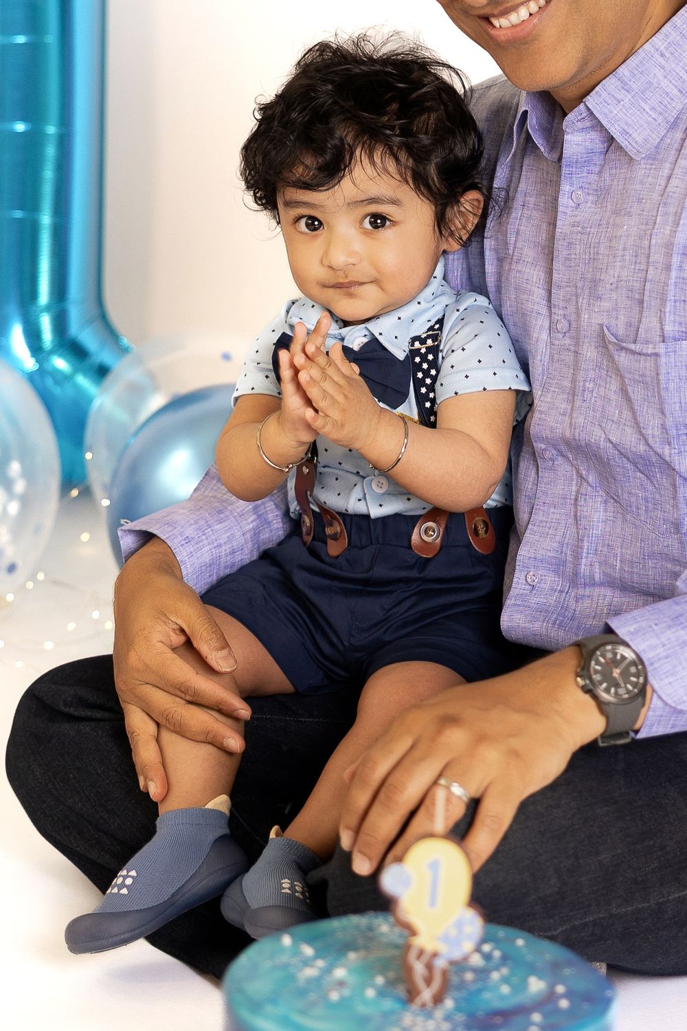 Toddler boy in suspenders clapping hands sits on father's lap at first birthday celebration with blue balloons.