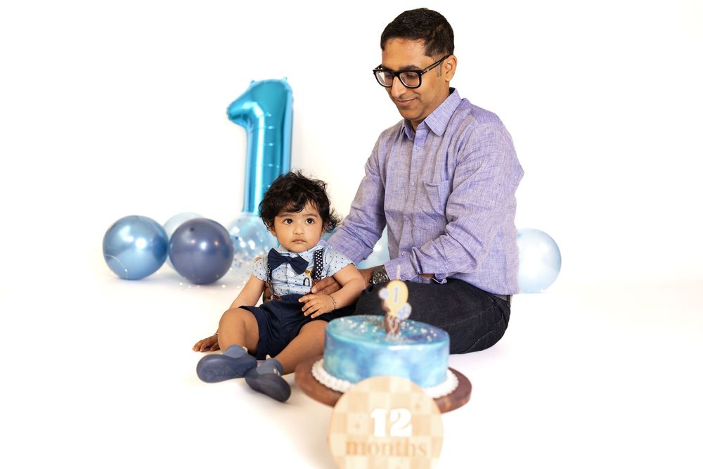 Father and baby boy celebrate 1st birthday with blue balloons and cake on white background.