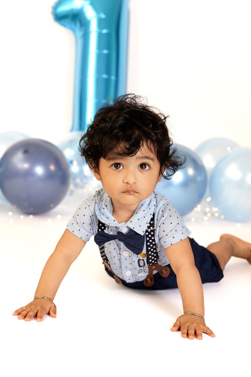 A baby boy in a blue bow tie outfit sits among blue balloons and a number one balloon for his first birthday.