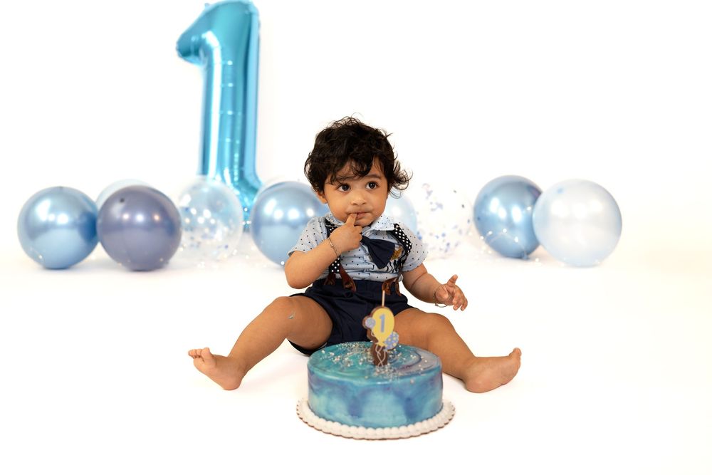 Baby boy smashing blue first birthday cake surrounded by blue balloons and a large number one balloon.