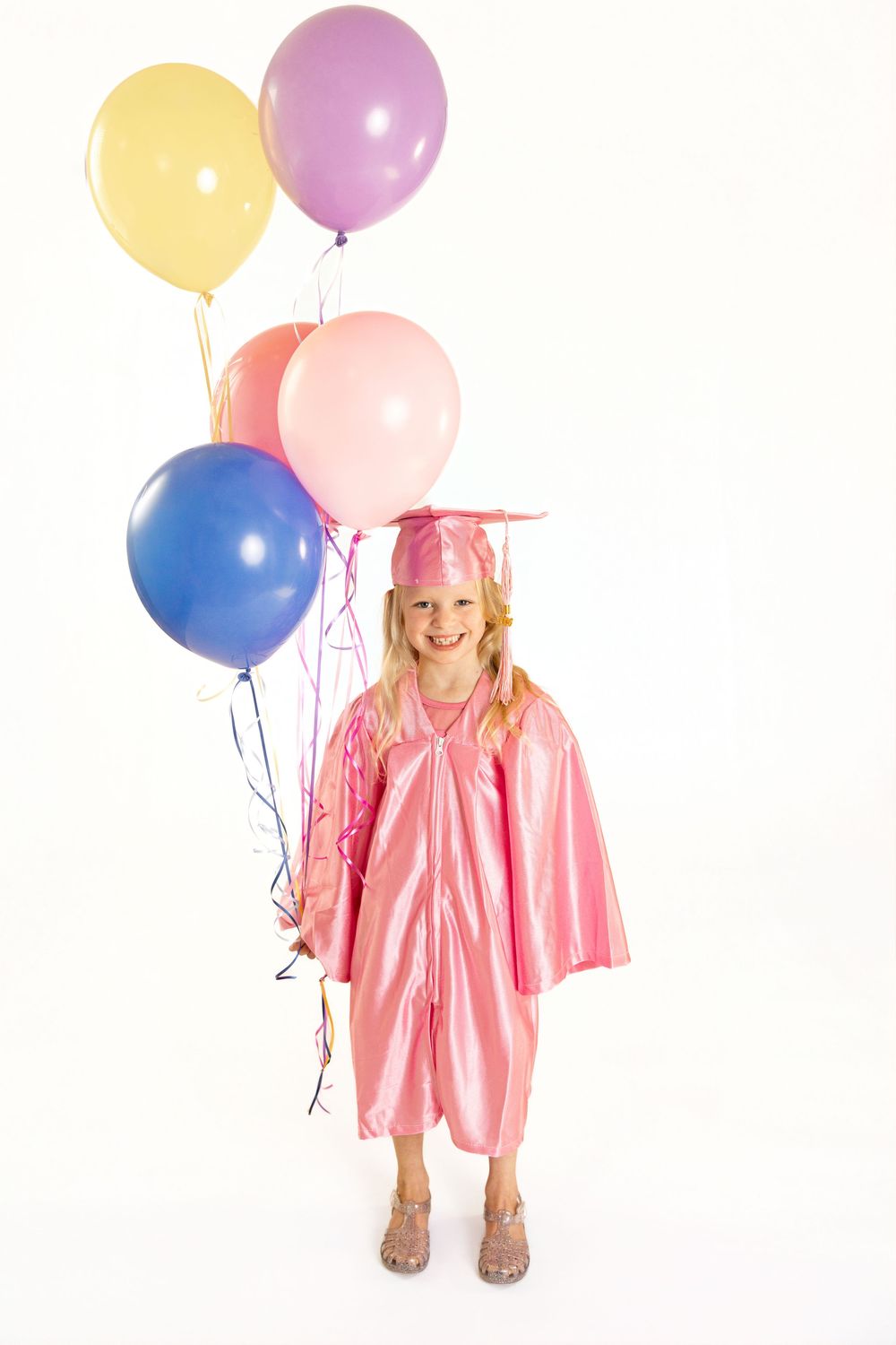 Young girl in pink graduation cap and gown smiling while holding colorful balloons on white background.