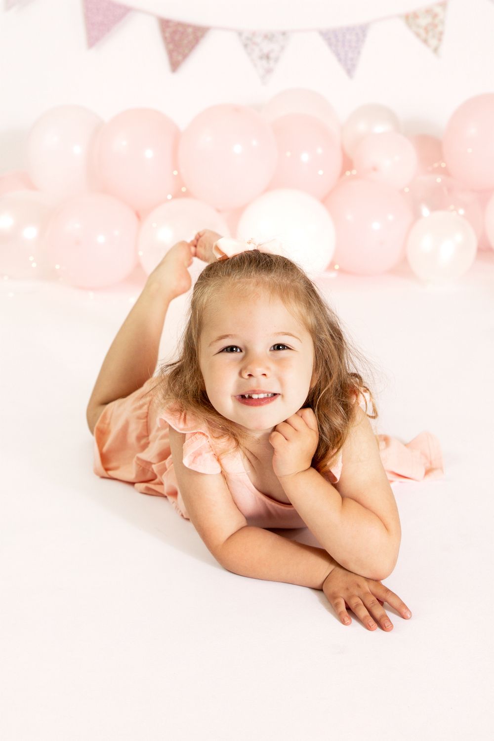 Smiling toddler girl in pink dress lying on white floor with pink balloons and bunting in background.