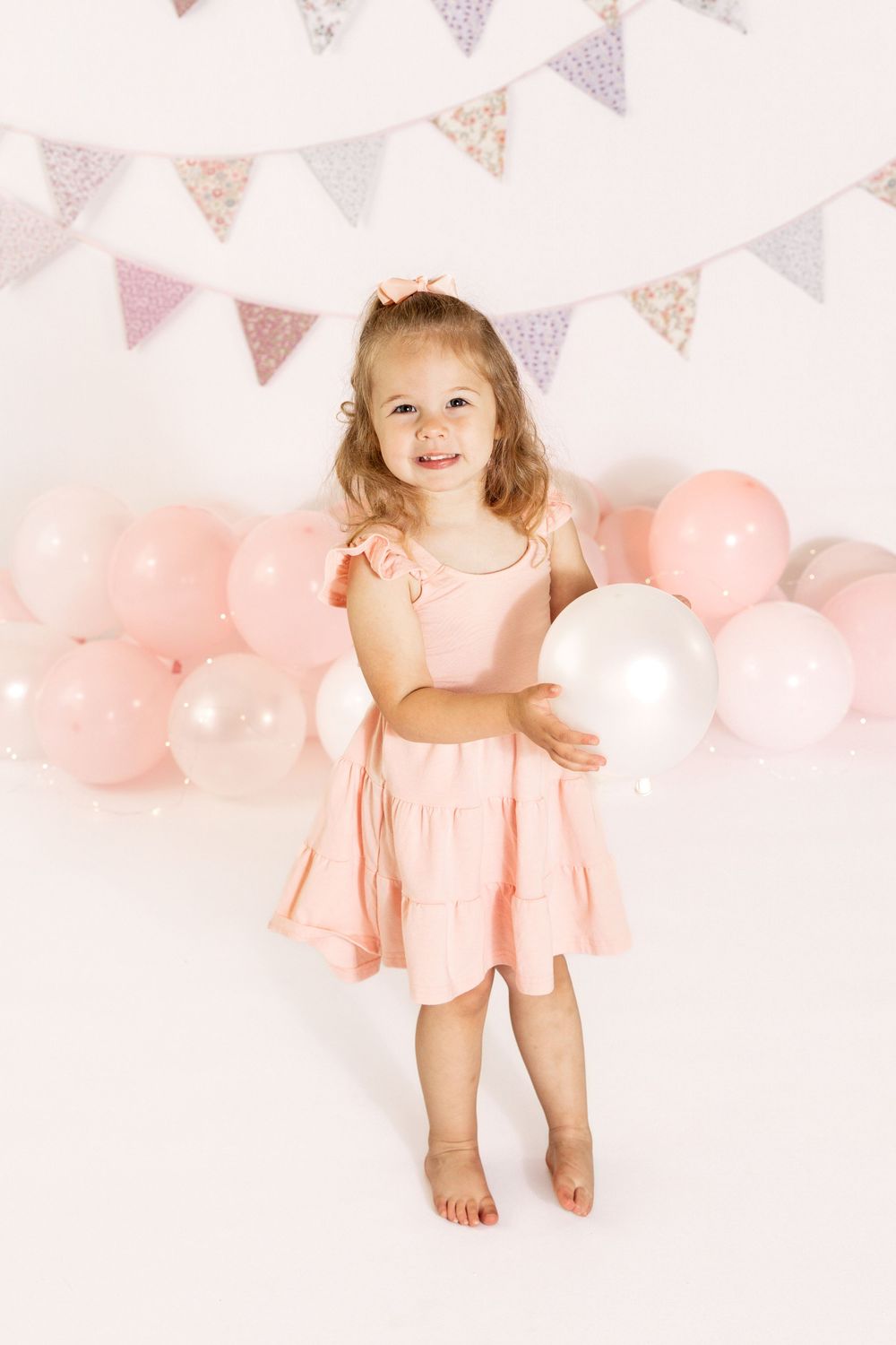 Smiling toddler girl in pink dress holding white balloon at birthday party with pink balloons and bunting backdrop.