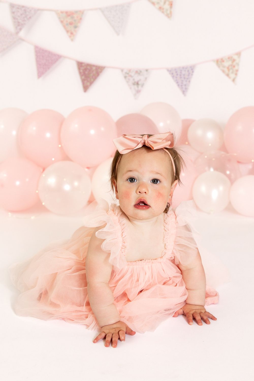Baby girl in pink tutu dress with bow headband sits before pink balloons and bunting at first birthday celebration.