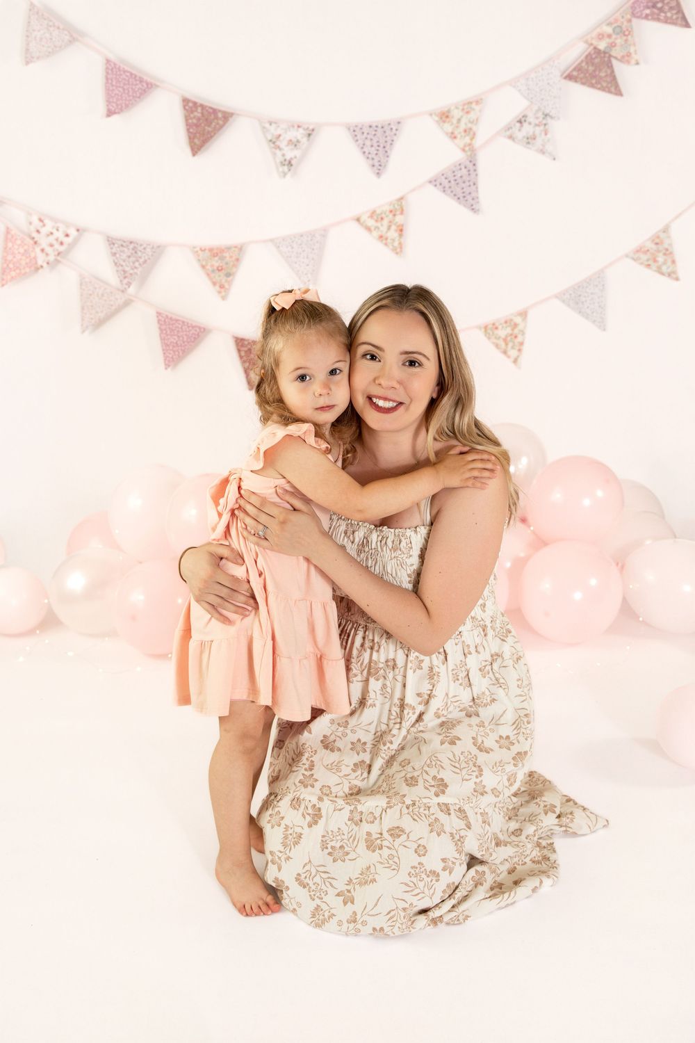 A mother hugs her toddler daughter in pink dress, posing before pastel balloons and bunting decorations.